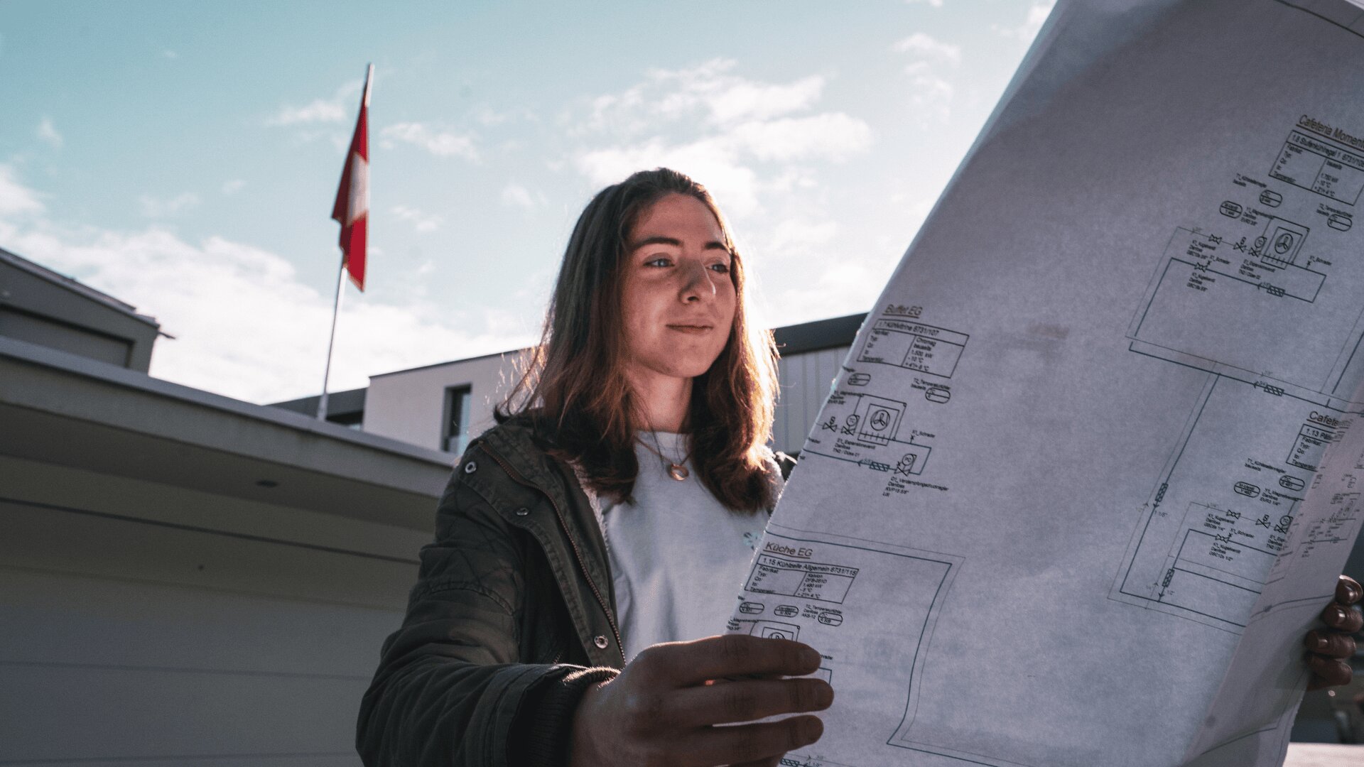 Eine junge Frau steht draußen und betrachtet große architektonische Entwürfe. Im Hintergrund sind bei teilweise bewölktem Himmel ein Gebäude und ein Fahnenmast mit einer rot-weißen Flagge zu sehen.