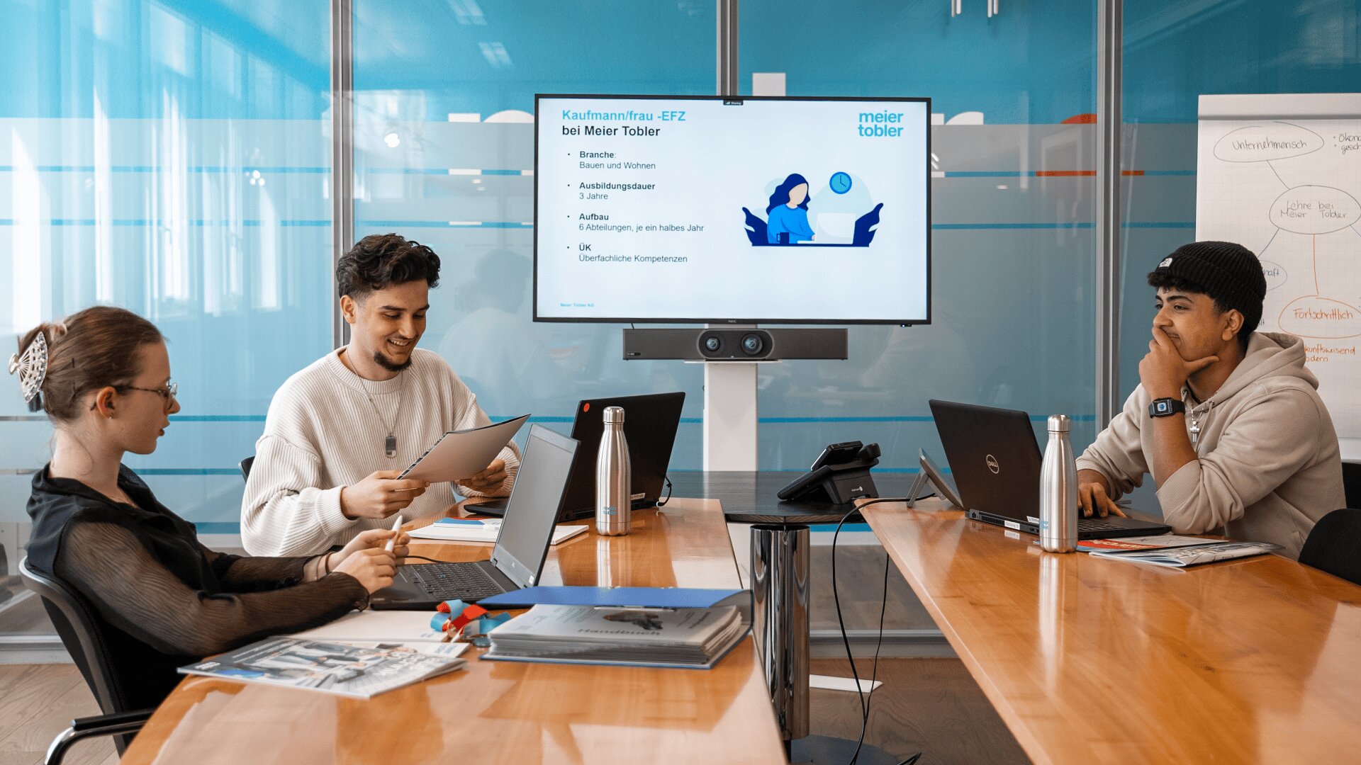Drei junge Erwachsene sitzen mit Laptops und Papieren an einem Konferenztisch und diskutieren in einem modernen Büro. Auf einem Bildschirm läuft eine Präsentation mit dem Titel "Kauffmannfrau EFZ bei Meier Tobler". Im Hintergrund steht ein Flipchart.