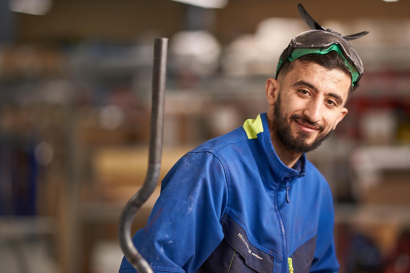 Ein Mann mit blauer Arbeitsjacke und Schutzbrille auf dem Kopf lächelt in einer Industrie- oder Werkstatteinrichtung, mit unscharfen Geräten und Regalen im Hintergrund.