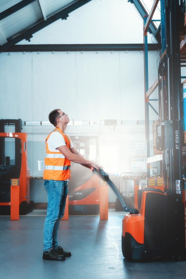 Ein Mann in einer Warnweste bedient einen orangefarbenen Hubwagen in einem Lagerhaus und schaut auf die Regale. Das Lager ist gut beleuchtet, im Hintergrund sind mehrere Gabelstapler und Regale zu sehen.