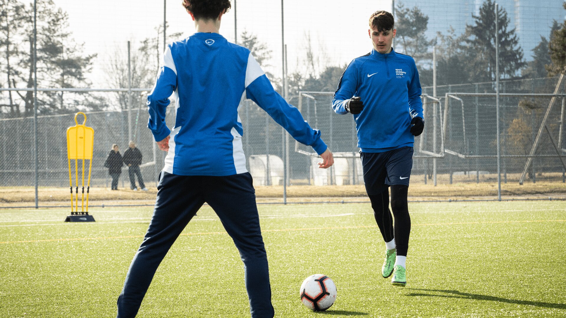 Zwei junge Männer in blauer Sportkleidung üben auf einem Außenplatz Fußball. Einer kontrolliert einen Ball, während der andere ihm gegenübersteht. Eine gelbe Übungspuppe und zwei Personen sind im Hintergrund in der Nähe eines Zauns zu sehen.