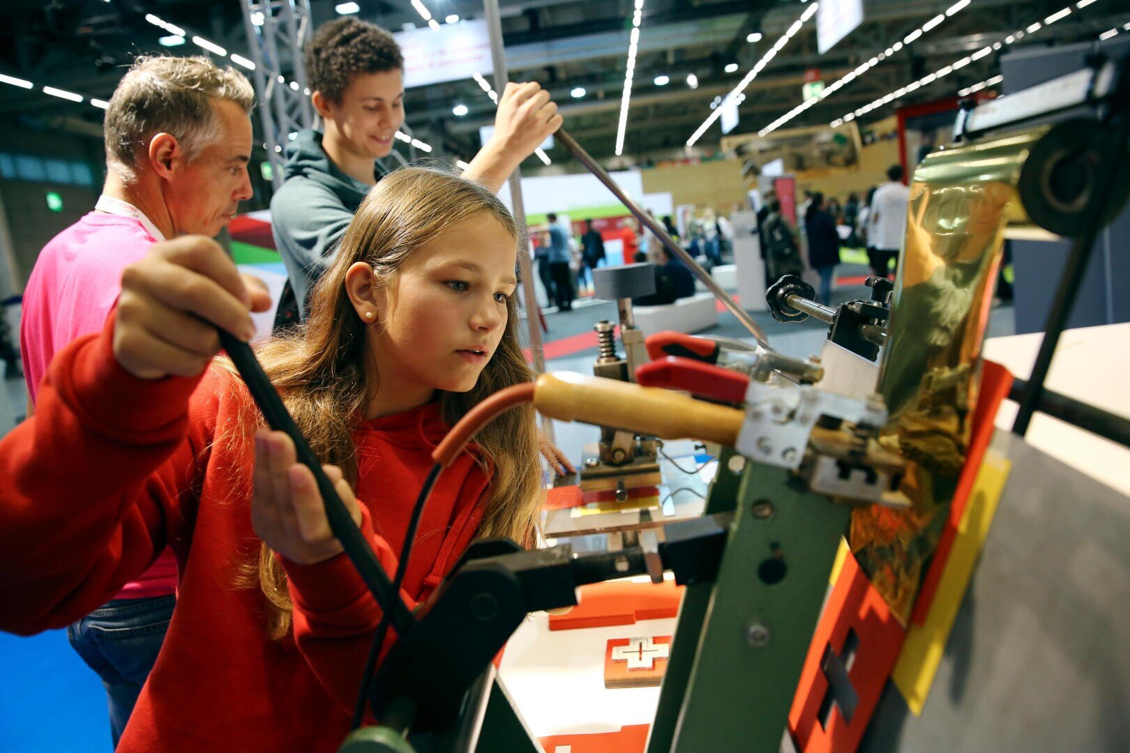 Ein Mädchen in einem roten Sweatshirt bedient eine Maschine in einer Ausstellung, während zwei Personen im Hintergrund zusehen und mit anderen Geräten interagieren. Die Umgebung ist hell und geschäftig, verschiedene Displays und Lichter sind zu sehen.