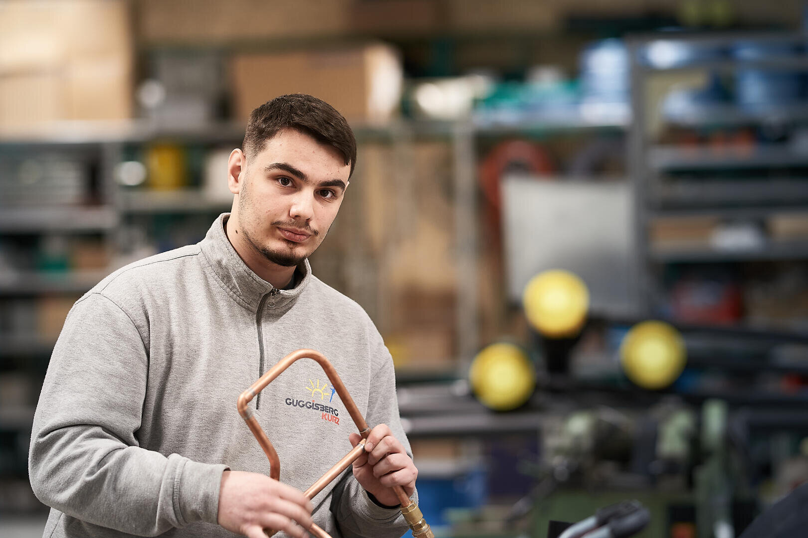 Ein Mann in einem grauen Sweatshirt hält ein Kupferrohr in einer mit Werkzeugen und Regalen ausgestatteten Werkstatt. Er blickt mit neutralem Blick in die Kamera. Der Hintergrund ist leicht unscharf und hebt den Mann und das Rohr hervor.