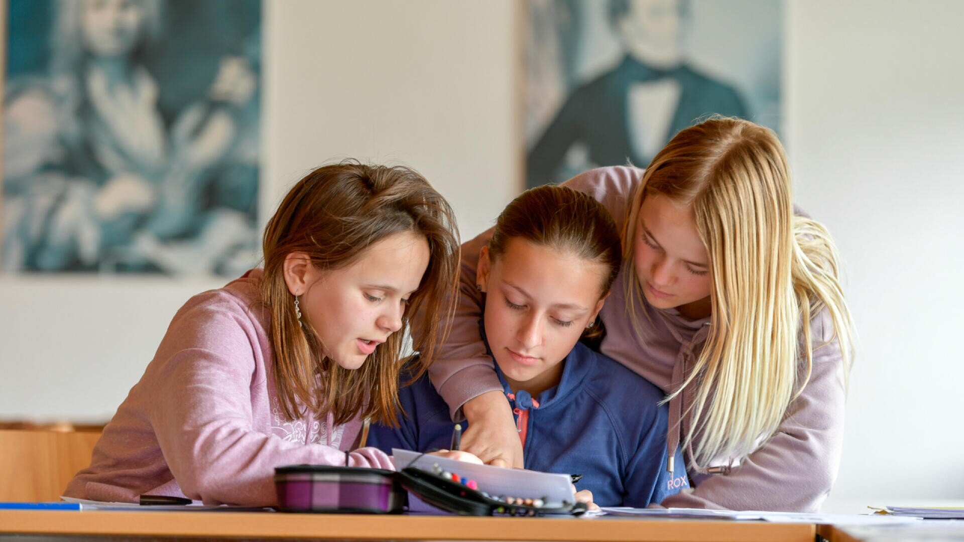 Drei junge Mädchen sitzen eng beieinander an einem Schreibtisch, arbeiten an Schulaufgaben und teilen sich ein Heft in einem Klassenzimmer. Zwei der Mädchen helfen dem Mädchen in der Mitte. An der Wand im Hintergrund hängen unscharfe Porträts.