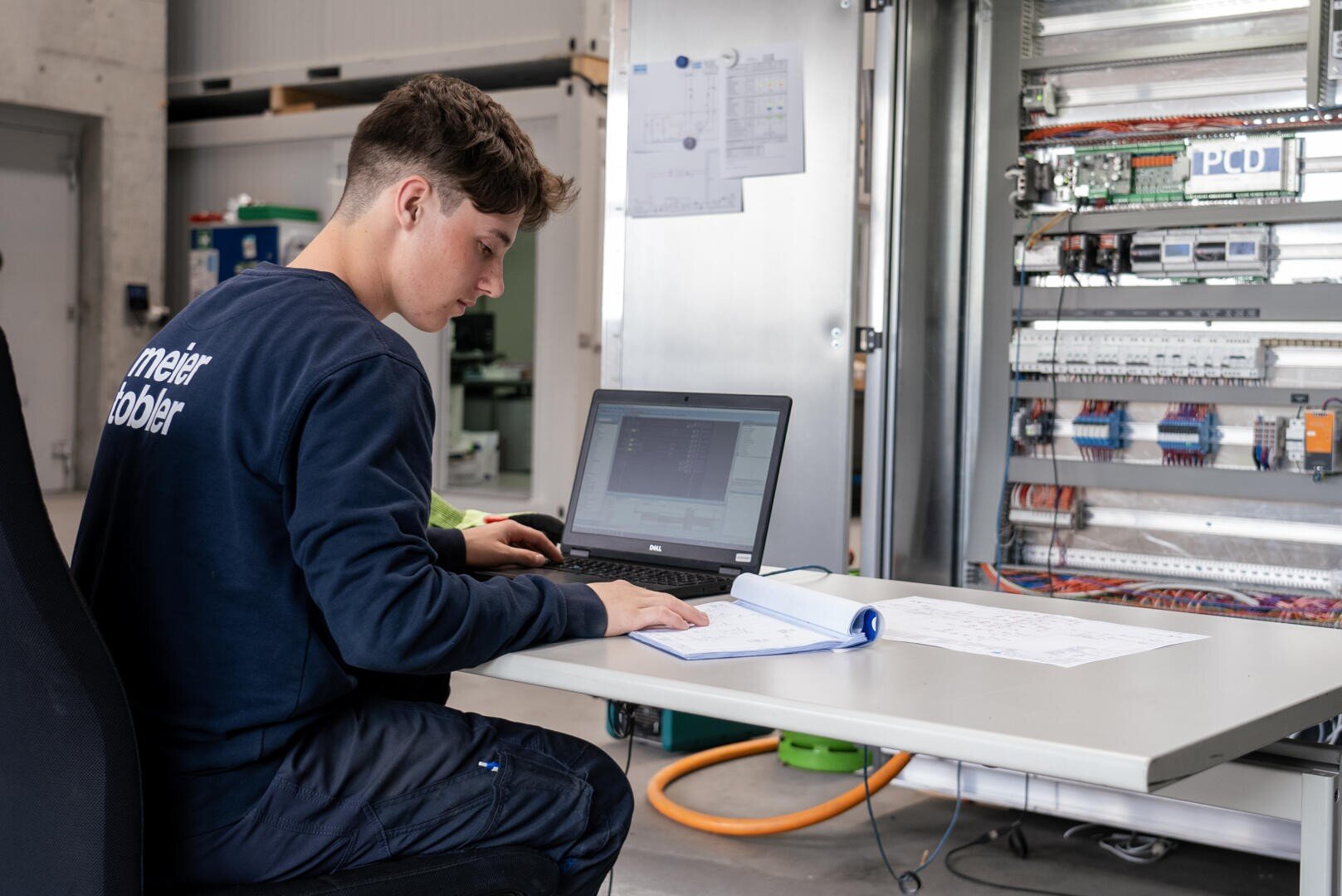 Ein junger Mann in Arbeitskleidung sitzt an einem Schreibtisch mit einem Laptop und einem aufgeschlagenen Handbuch vor einem offenen elektrischen Schaltschrank und scheint sich auf seine technische Arbeit zu konzentrieren.