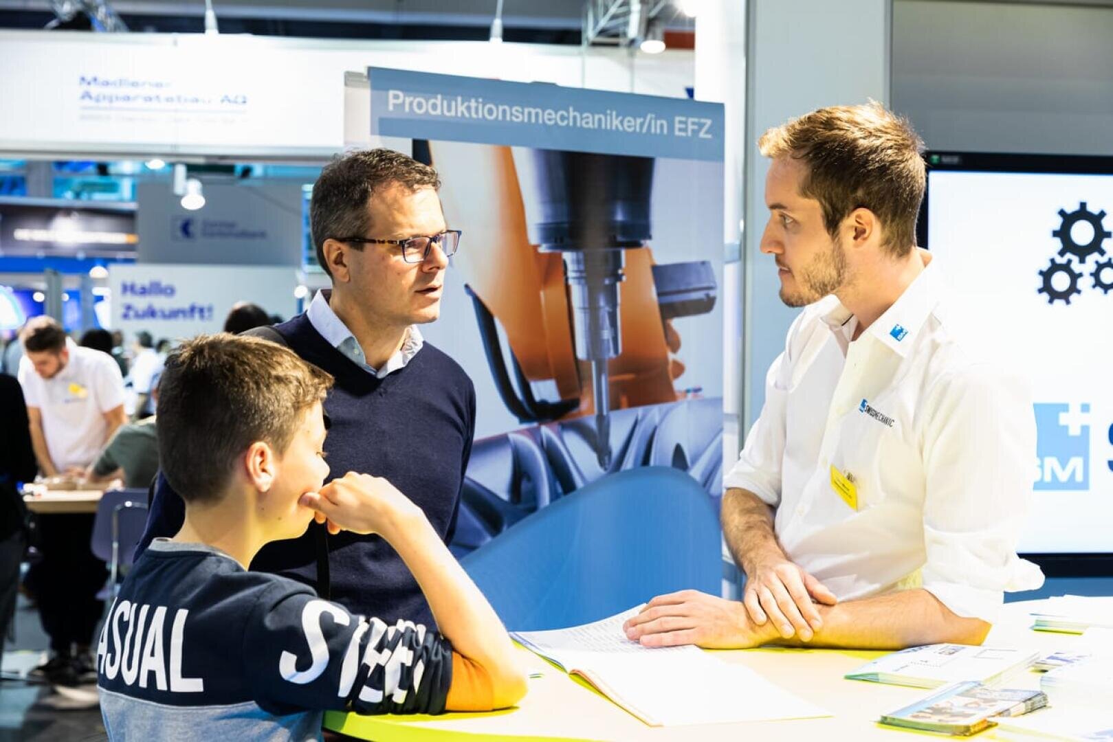 Ein Mann und ein Junge sprechen mit einem Vertreter an einem Informationsstand über Industriemechanik. Sie unterhalten sich, während im Hintergrund Lehrplakate und Bilder von Maschinen zu sehen sind.