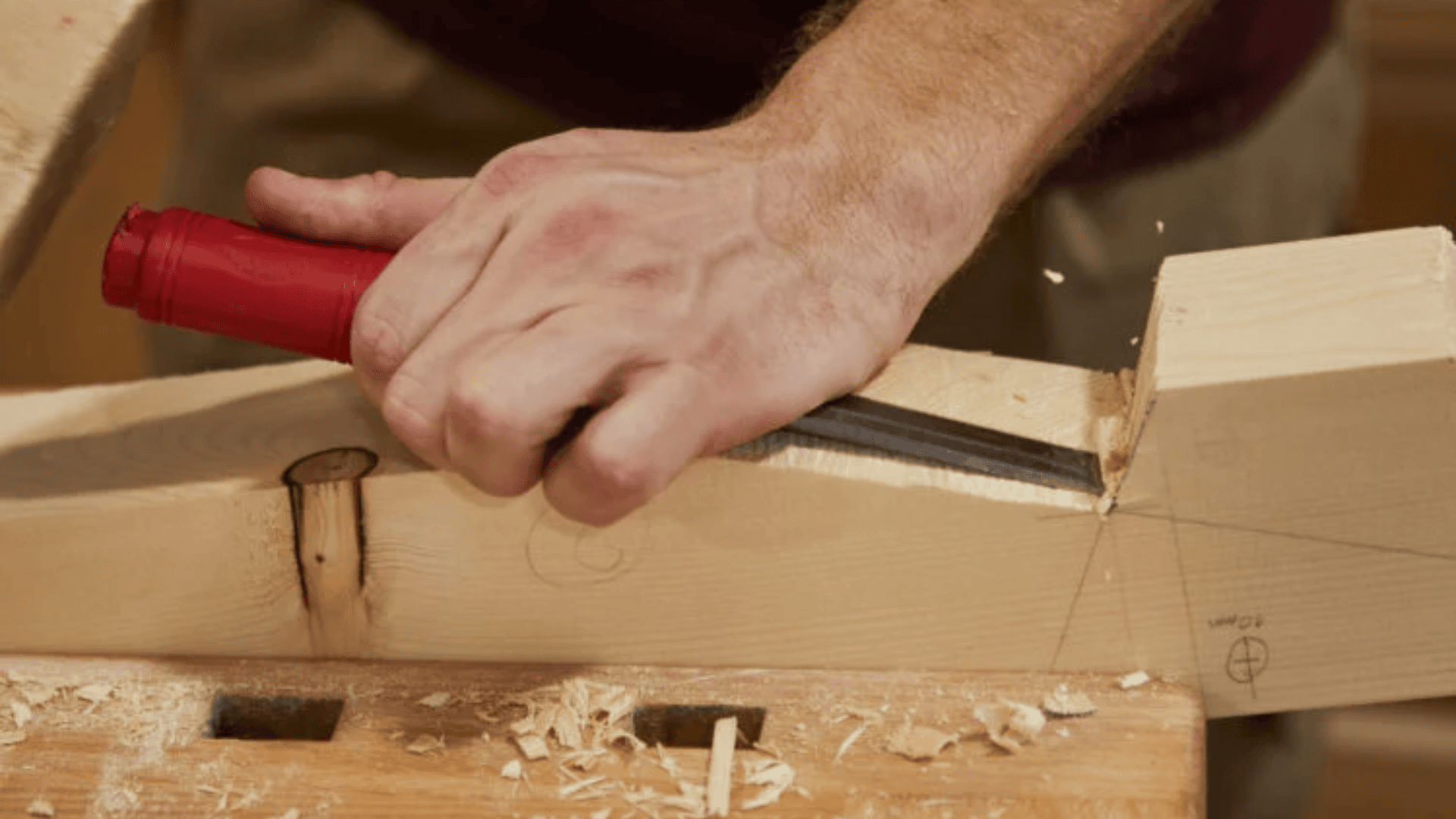Nahaufnahme der Hand einer Person, die mit einem Meißel und einem Hammer mit rotem Griff eine Kerbe in ein Stück helles Holz auf einer Werkbank schnitzt, wobei Holzspäne verstreut sind.