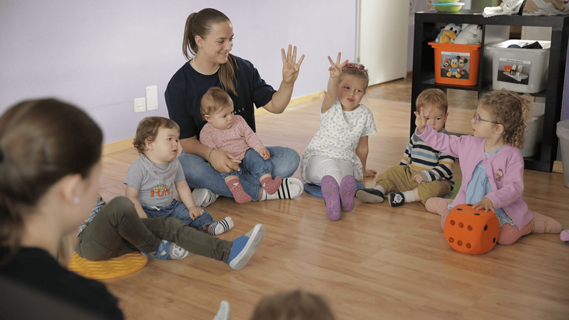 Eine Gruppe kleiner Kinder sitzt mit einer Lehrerin in einem Kreis auf dem Boden eines Klassenzimmers. Ein Kind hält seine Hand hoch, während ein anderes einen orangefarbenen Schaumstoffwürfel hält. Die Atmosphäre wirkt spielerisch und lehrreich.