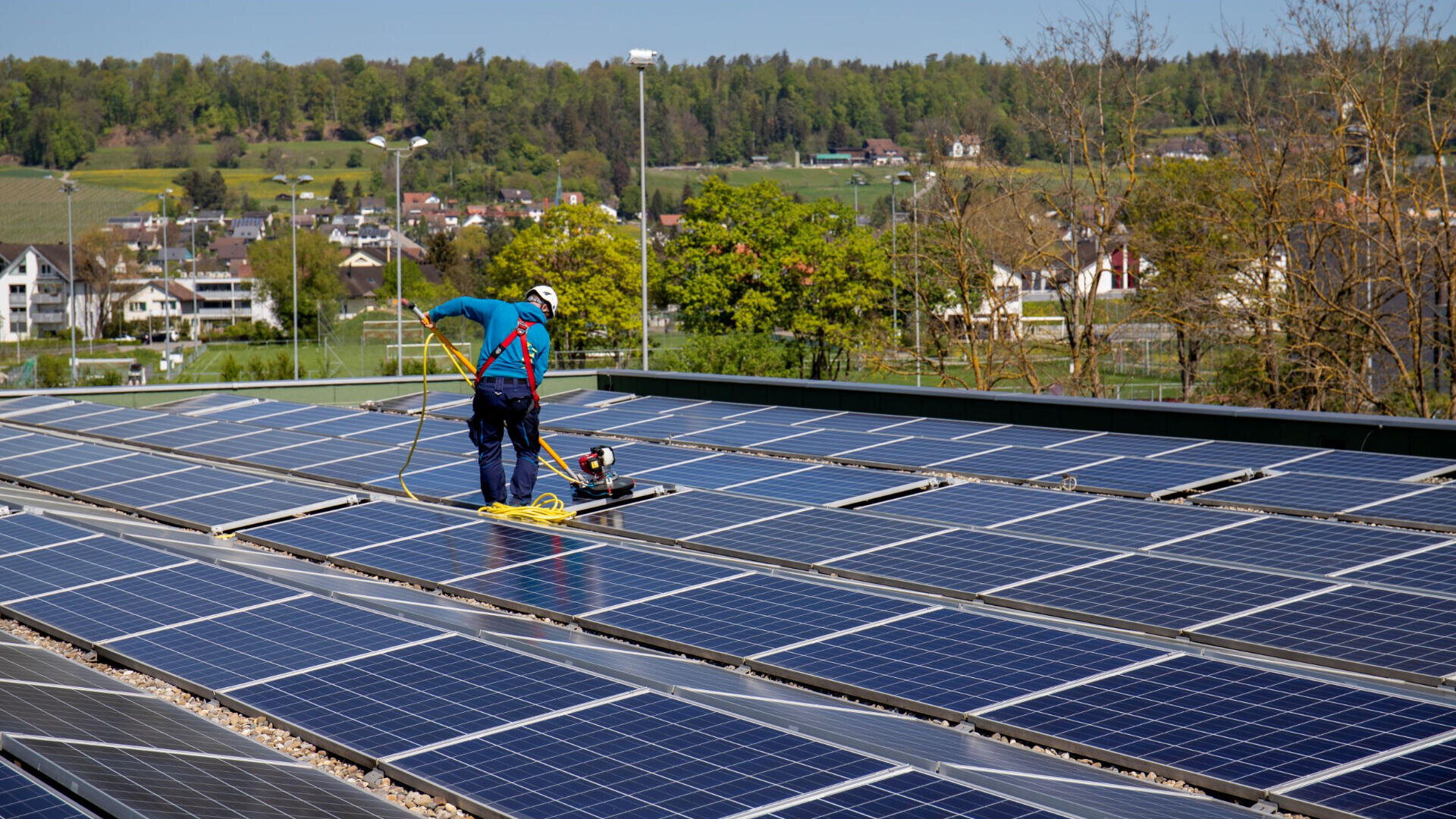 Ein Arbeiter in Schutzkleidung reinigt mit einer langen Bürste und einem Schlauch Solarzellen auf einem Dach. Im Hintergrund sind bei klarem Himmel Bäume und Häuser zu sehen.