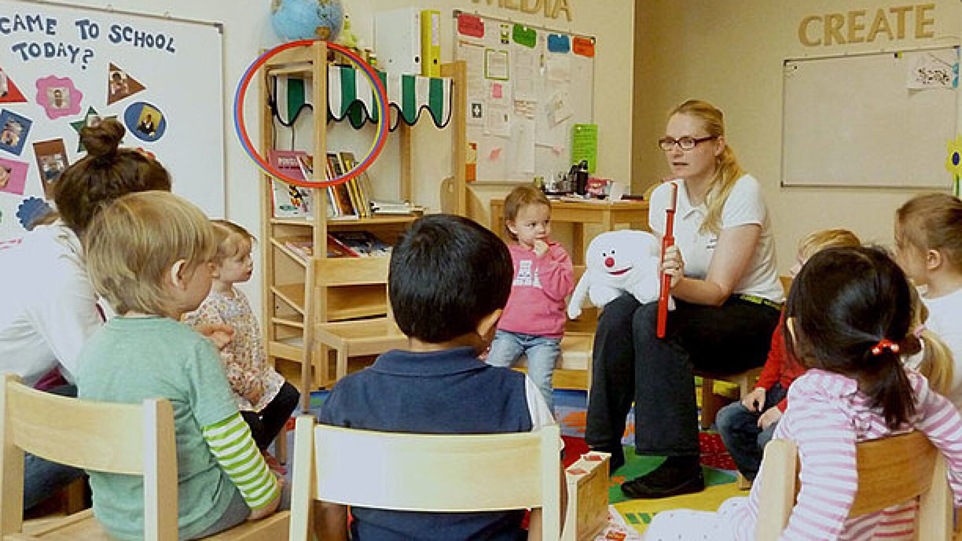 Eine Lehrerin sitzt mit kleinen Kindern in einem Klassenzimmer im Kreis, hält eine Handpuppe und einen roten Stock, während die Kinder aufmerksam zuhören und zusehen. Die Dekoration des Klassenzimmers und die Lernmaterialien sind sichtbar.
