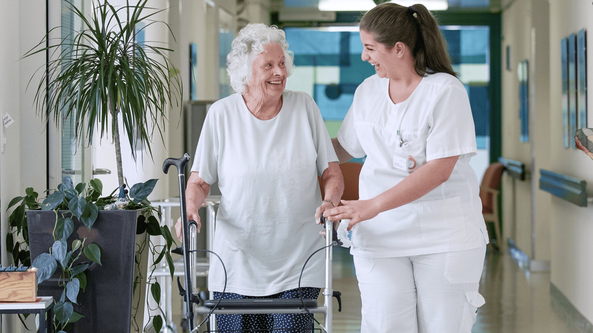 Eine ältere Frau, die eine Gehhilfe benutzt, lächelt, während sie von einer Krankenschwester in weißem Kittel unterstützt wird, während sie einen hellen Flur mit Grünpflanzen in der Nähe entlanggehen.