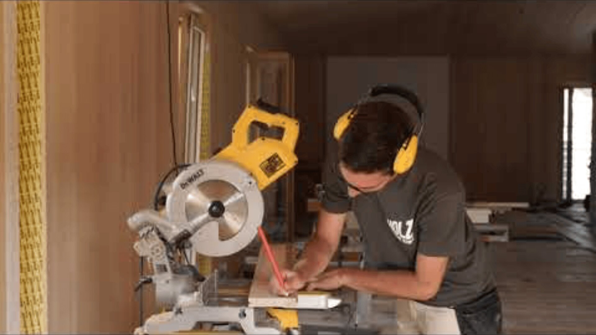 Eine Person, die einen Gehörschutz trägt, markiert ein Holzstück mit einem Bleistift und bereitet sich darauf vor, es mit einer gelben Gehrungssäge in einem im Bau befindlichen Raum zu schneiden.