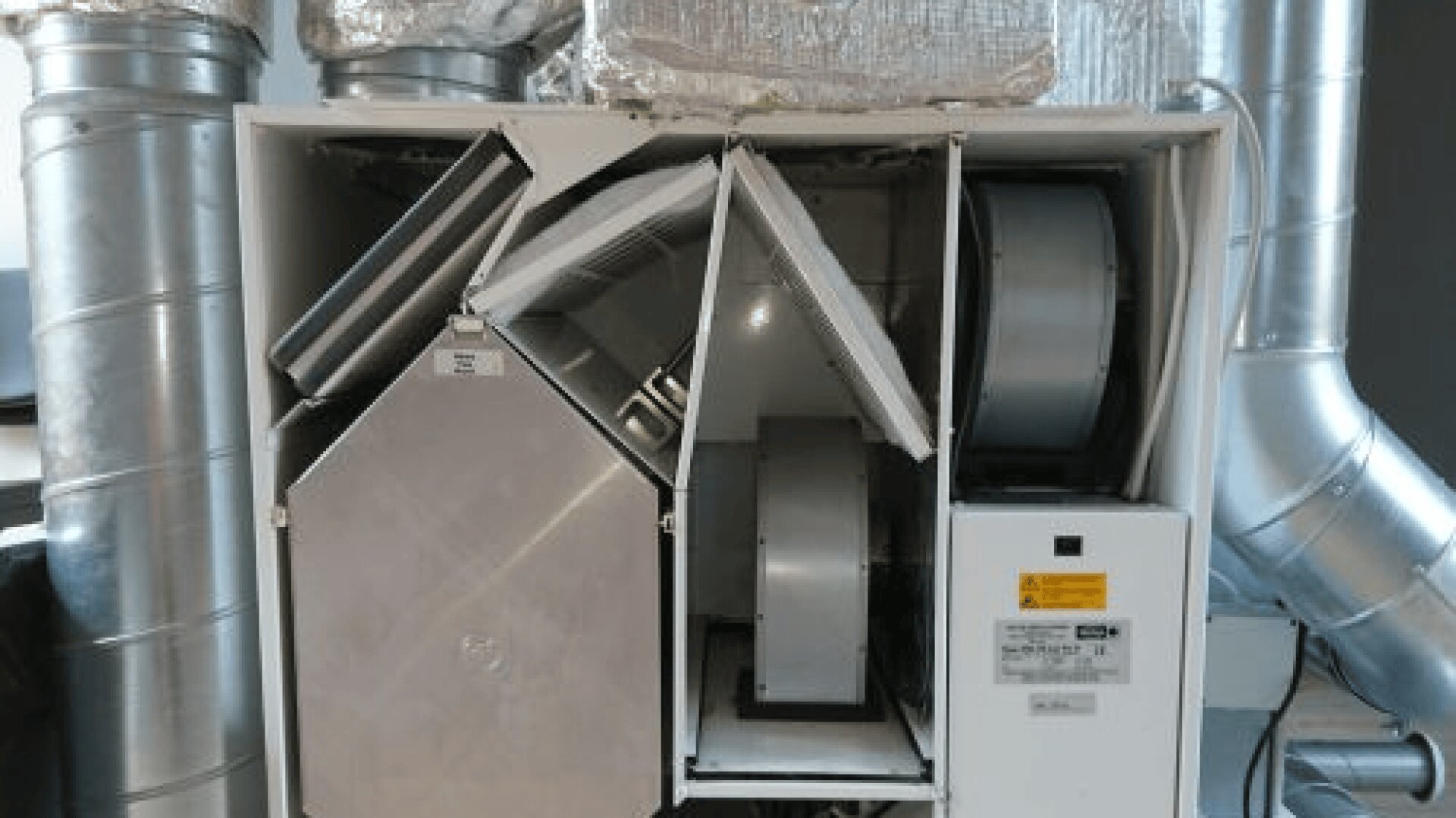 Ein offenes industrielles HLK-Gerät mit internen Komponenten wie Ventilatoren, Filtern und Kanälen, wobei im Hintergrund Metallrohre und Isolierung zu sehen sind.