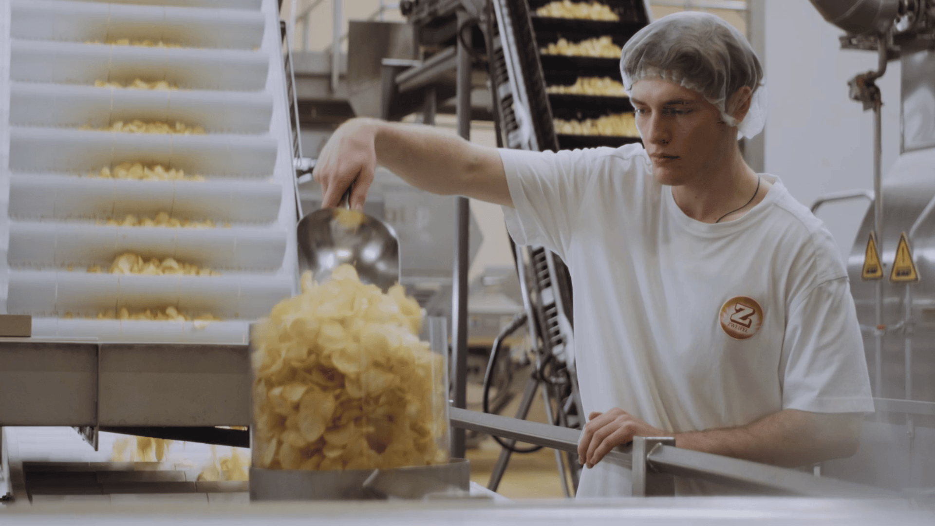 Ein Arbeiter mit Haarnetz und weißem Hemd schüttet in einer Snackfood-Fabrik Kartoffelchips aus einer Metallschaufel in einen Behälter, im Hintergrund sind Förderbänder und Geräte zu sehen.
