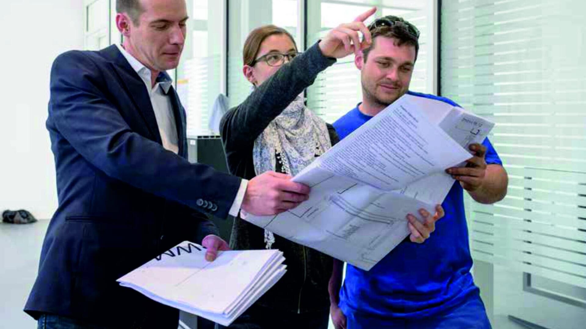 Drei Personen stehen in einem Haus und diskutieren über Architekturpläne. Eine Person in einem Anzug hält mehrere Dokumente in der Hand, während eine andere Person gestikuliert und nach vorne zeigt. Die dritte Person im blauen Hemd schaut sich die Papiere aufmerksam an.