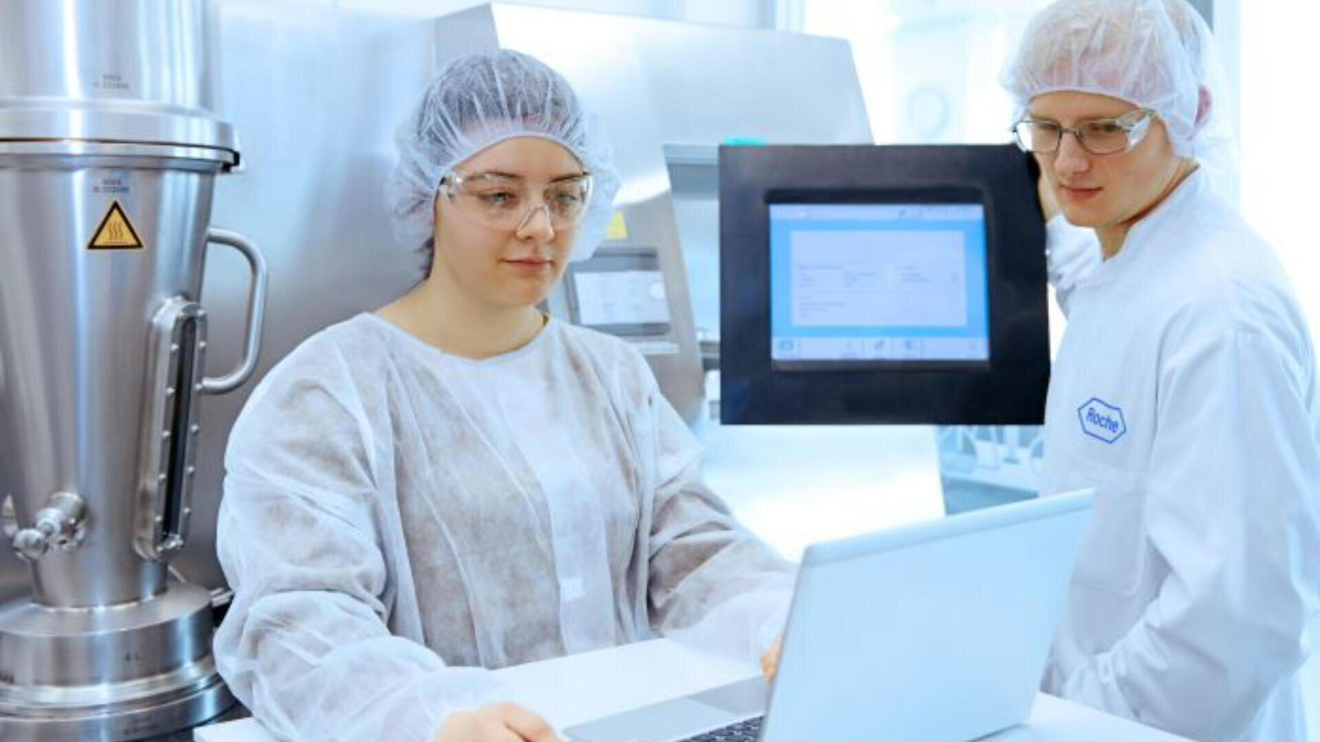 Zwei Personen in Reinraumanzügen und Haarnetzen arbeiten in einem Labor. Einer benutzt einen Laptop, während der andere auf einen Monitor schaut. Im Hintergrund sind Laborgeräte aus rostfreiem Stahl zu sehen.