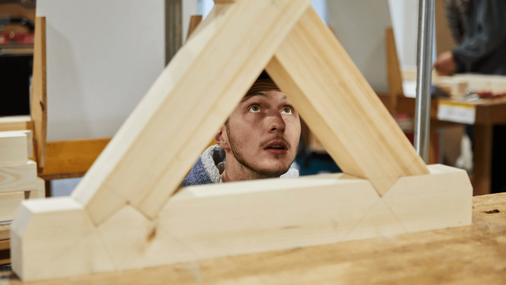Ein Mann blickt durch einen dreieckigen Holzrahmen in einer Werkstatt auf und konzentriert sich auf sein Schreinerprojekt.