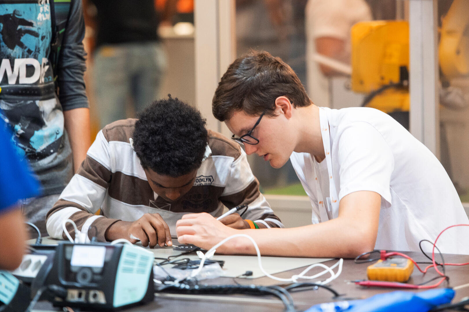 Zwei junge Männer untersuchen und bearbeiten eine elektronische Leiterplatte an einem Tisch, umgeben von Werkzeugen und Drähten, in einer kollaborativen, praxisorientierten Lernumgebung.