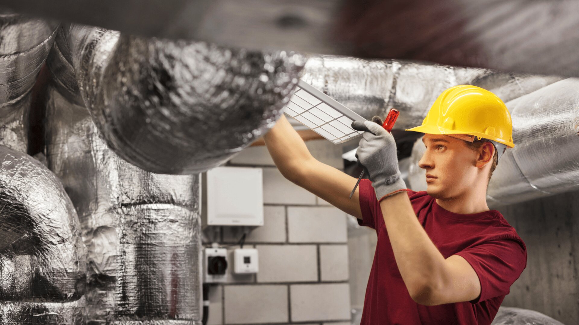 Ein Arbeiter, der einen gelben Schutzhelm und Handschuhe trägt, inspiziert oder repariert Luftkanäle in einer Industrieumgebung, umgeben von großen silbernen Lüftungsrohren.