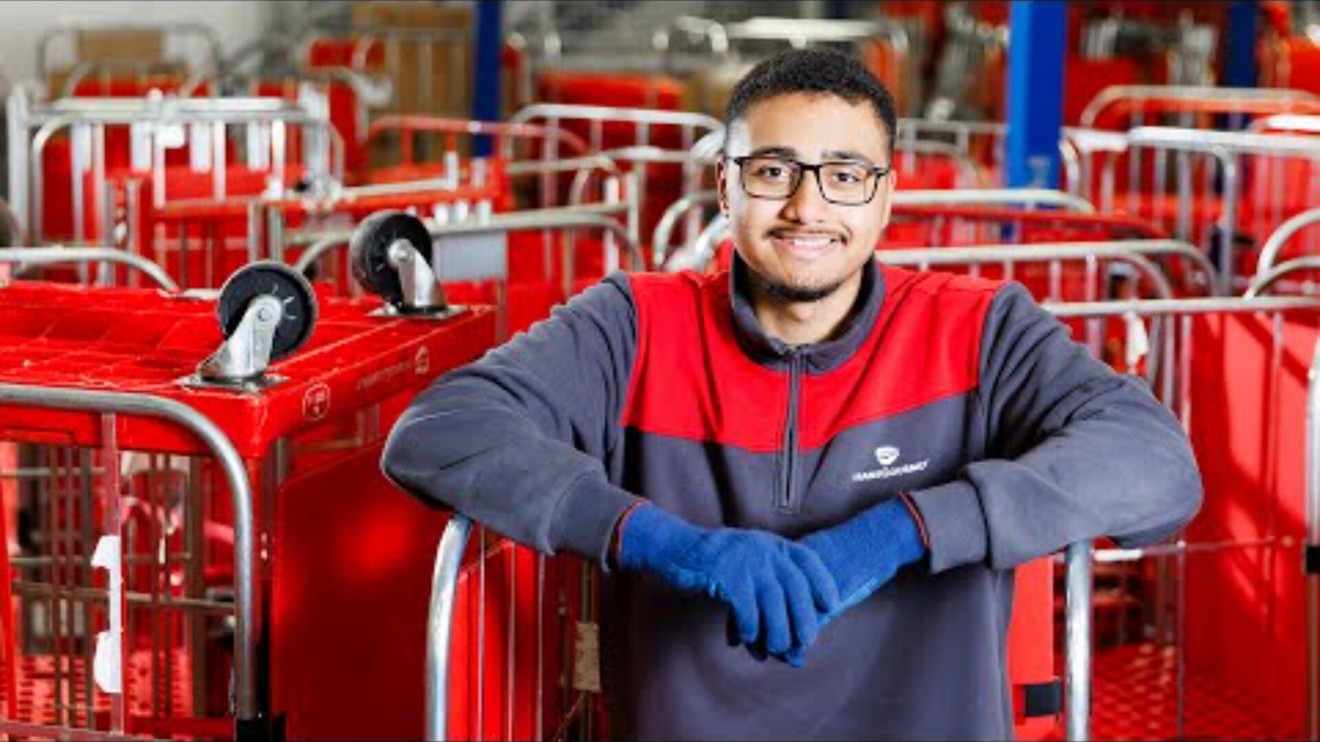 Ein Mann mit Brille, blauen Handschuhen und einer rot-grauen Uniform lächelt, während er sich auf einen roten Wagen in einem Lagerhaus stützt, das mit ähnlichen roten Wagen gefüllt ist.