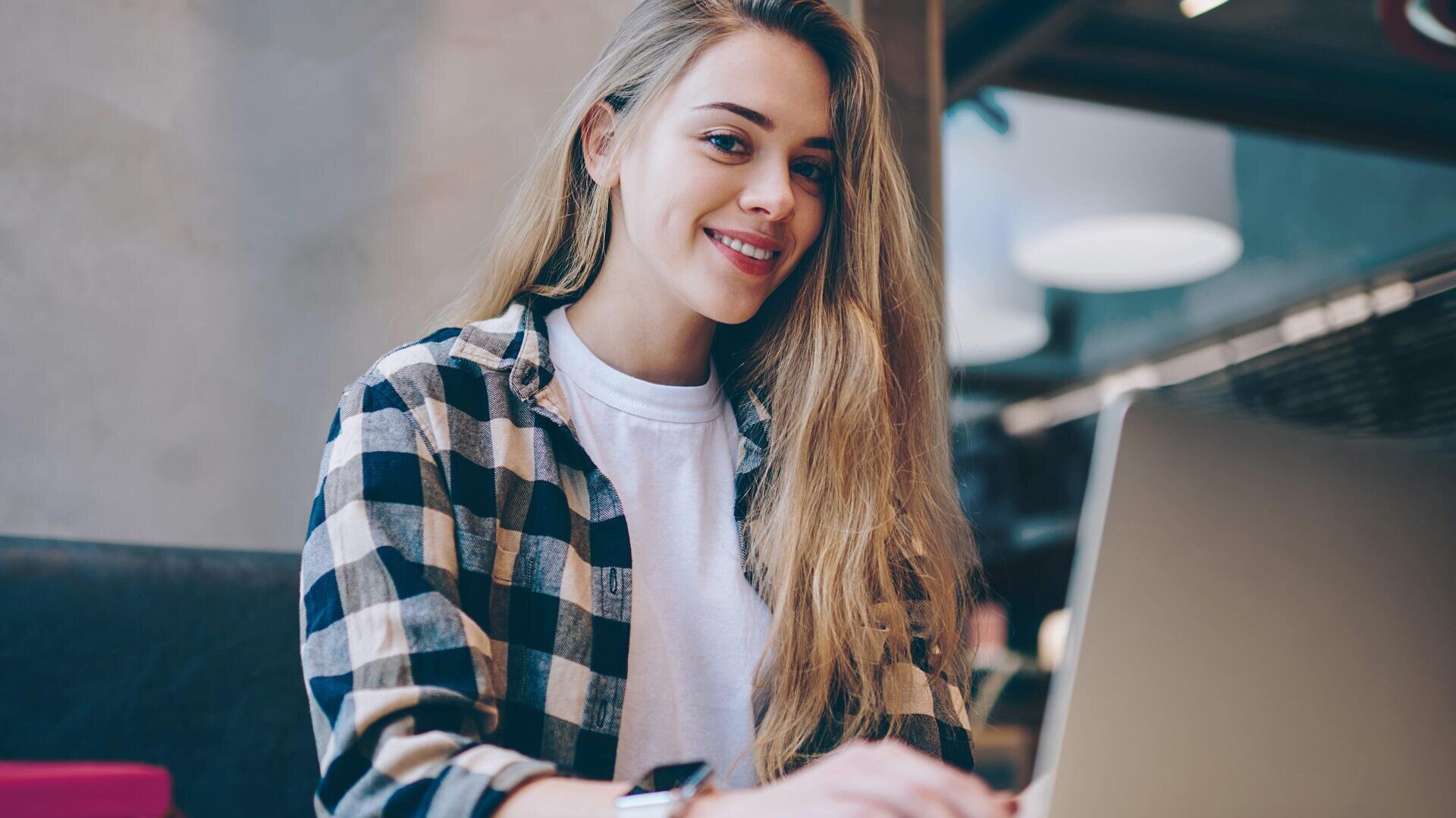 Eine junge Frau mit langen blonden Haaren, die ein schwarz-weiß kariertes Hemd und ein weißes T-Shirt trägt, sitzt in einem Haus und lächelt, während sie an einem Laptop arbeitet.