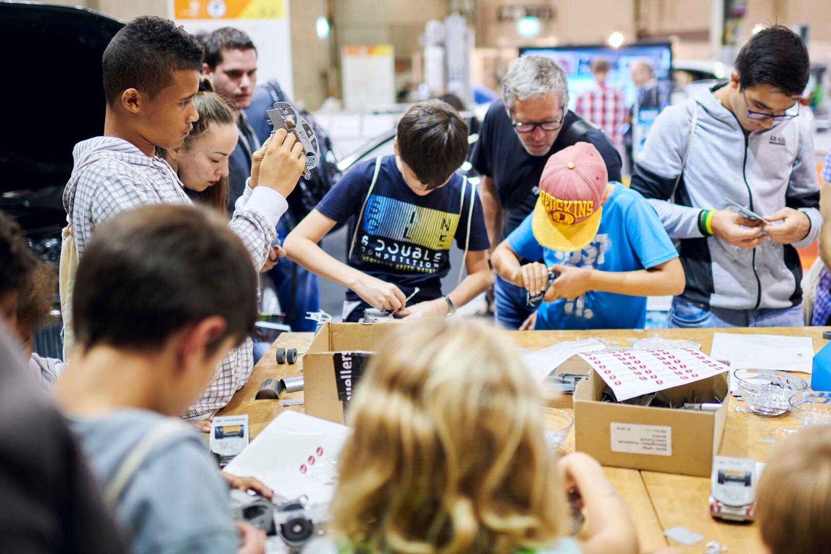 Eine Gruppe von Kindern und Erwachsenen versammelt sich um einen Tisch und baut kleine mechanische oder elektronische Bausätze zusammen. Sie konzentrieren sich auf ihre Projekte, die wie ein praktischer Workshop oder eine Wissenschaftsmesse aussehen.