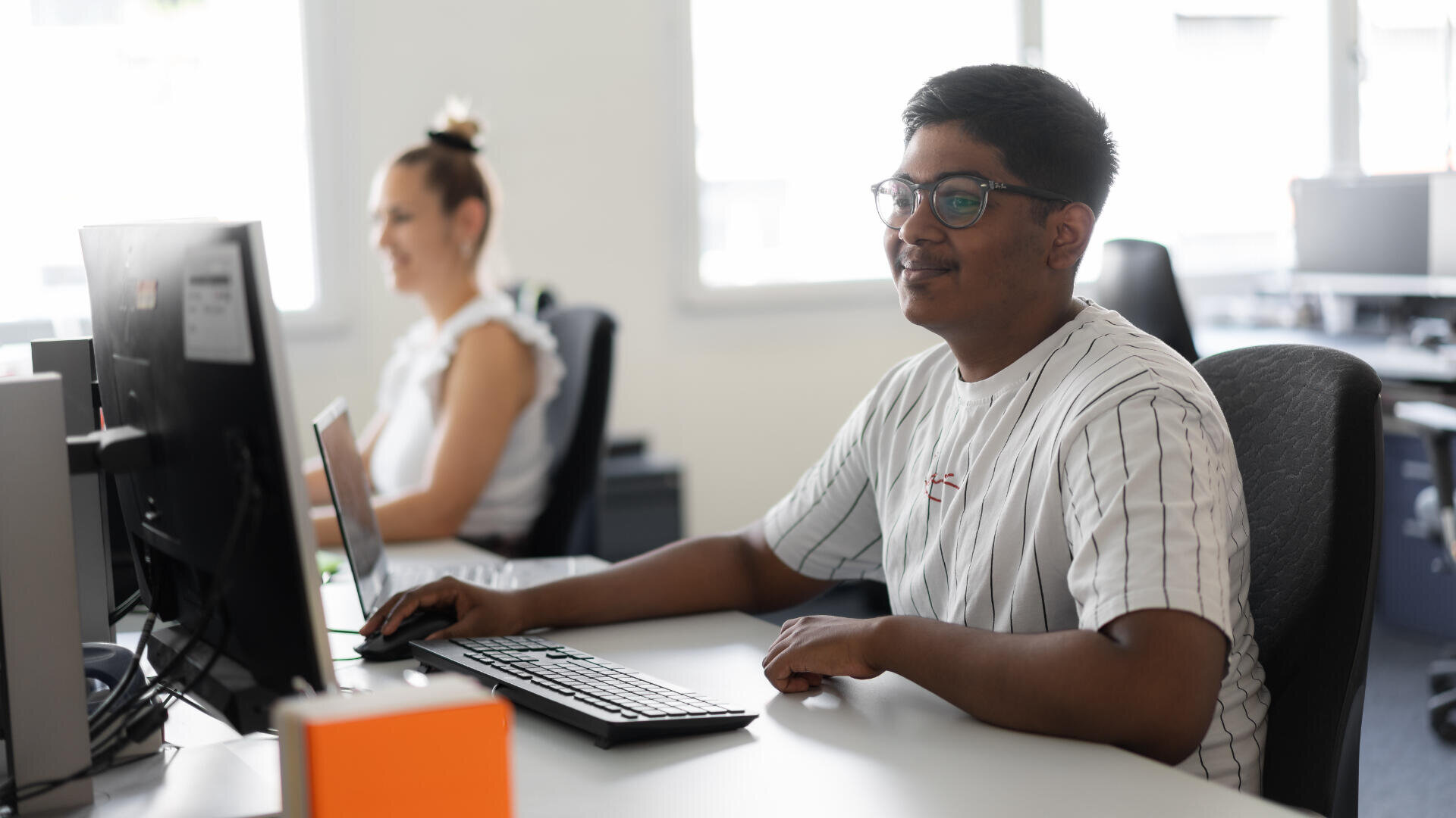 Ein Mann mit Brille sitzt an einem Schreibtisch mit einem Computer und lächelt in die Kamera. Im Hintergrund arbeitet eine Frau an einem anderen Schreibtisch ebenfalls an einem Computer in einem hellen, modernen Büro.