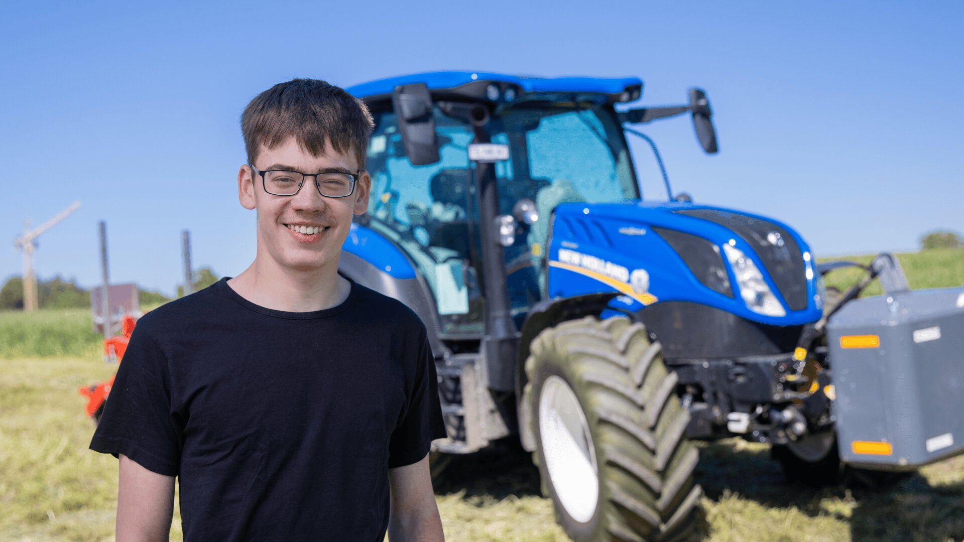 Ein junger Mann mit Brille und schwarzem T-Shirt steht lächelnd vor einem großen blauen Traktor auf einem sonnigen, grasbewachsenen Feld.