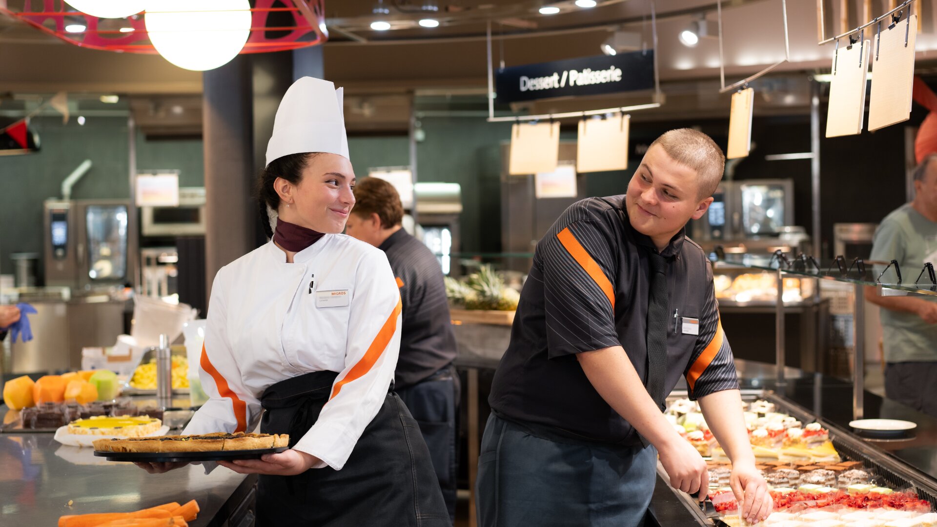Eine Köchin, die eine Torte in der Hand hält, und ein männlicher Angestellter, der das Essen zubereitet, lächeln gemeinsam hinter einer Buffet-Theke in einer gut beleuchteten, modernen Cafeteria, in der Desserts und Obst ausgestellt sind.