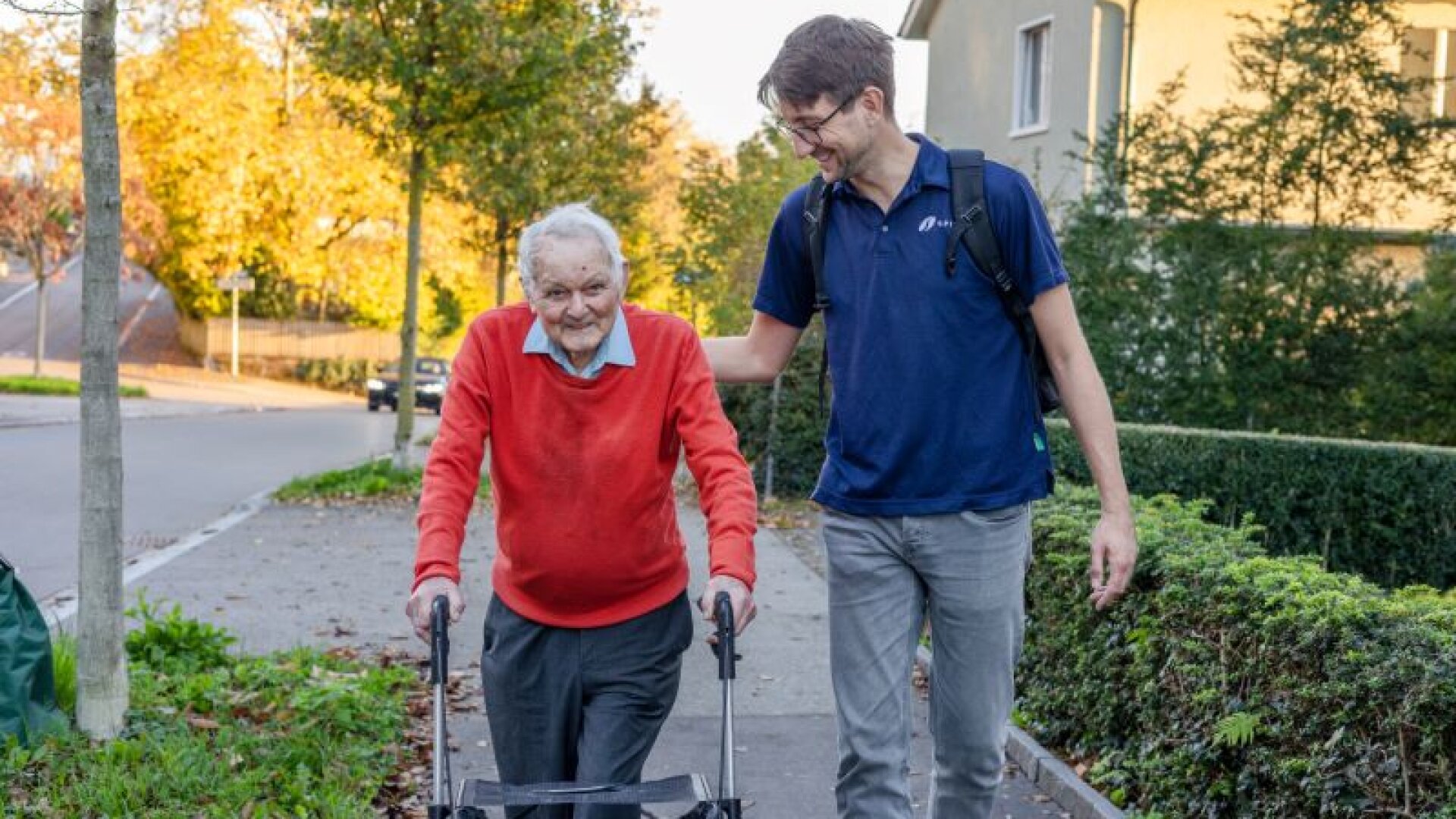 Ein älterer Mann, der eine Gehhilfe benutzt, lächelt, während er draußen auf einem Bürgersteig spazieren geht, begleitet von einem jüngeren Mann mit Brille, der ihn sanft stützt und lächelt. Im Hintergrund sind Bäume und Häuser zu sehen.