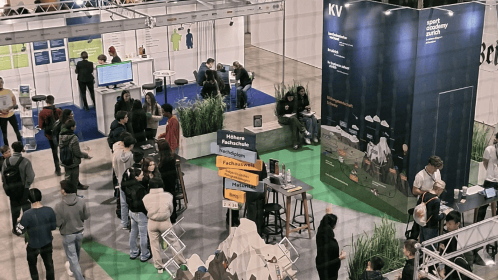 Ausstellungshalle mit Menschen, die verschiedene Stände besuchen, einschließlich eines Informationsbereichs mit Schildern und einem großen dunklen Stand auf der rechten Seite, umgeben von grünem und blauem Bodenbelag und Informationsdisplays.