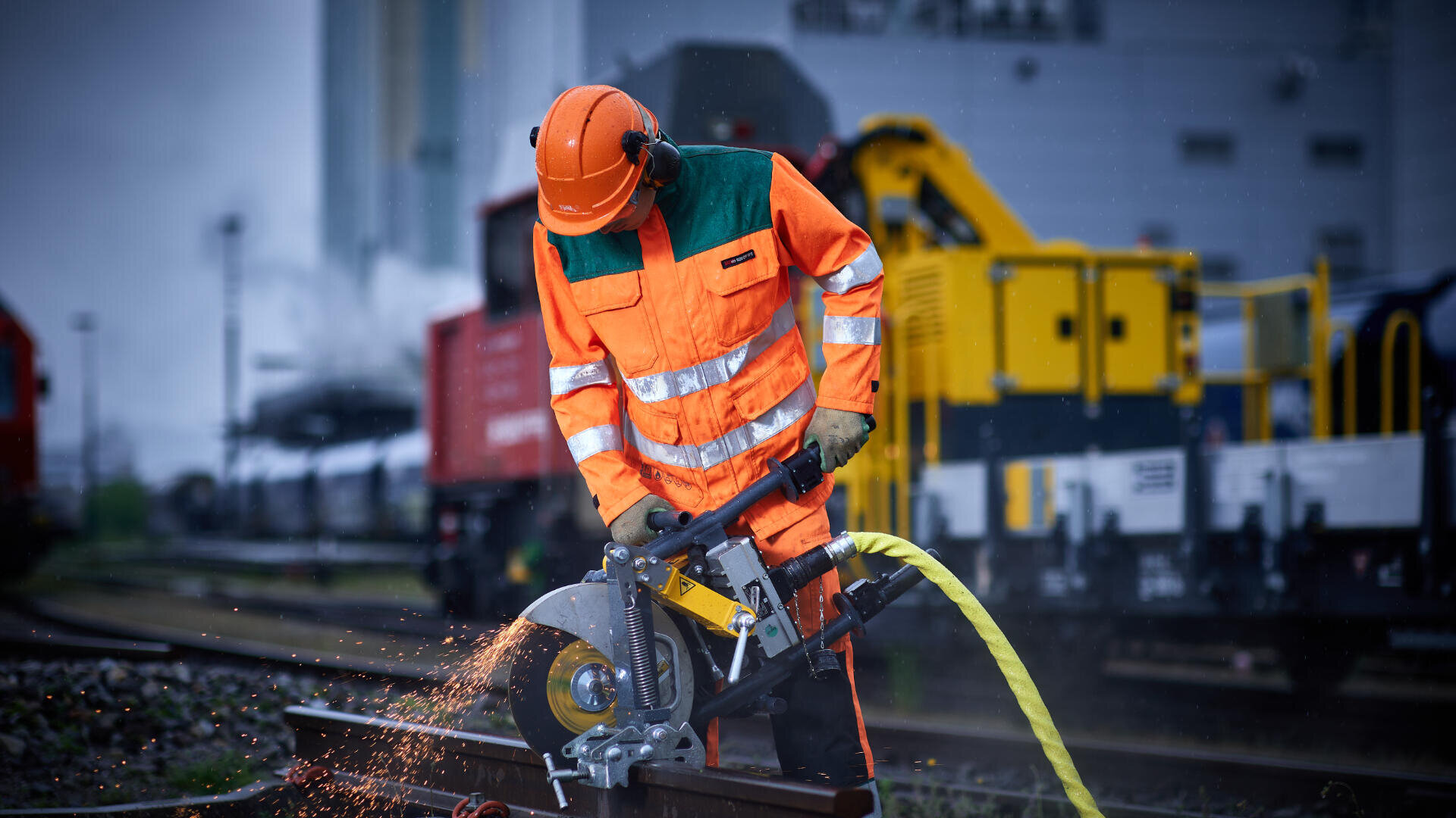 Ein Arbeiter in orangefarbener Warnkleidung und mit Helm schneidet mit einer Motorsäge eine Metallschiene und erzeugt dabei Funken, während im Hintergrund Industrieanlagen und Züge zu sehen sind.