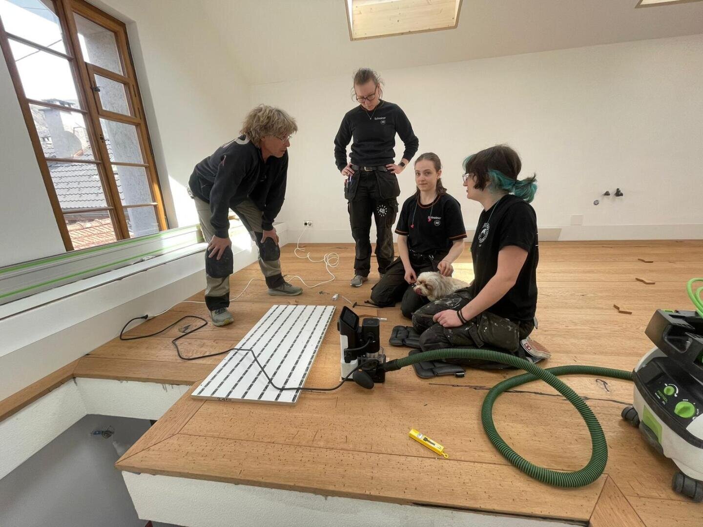 Vier Personen in Arbeitskleidung sitzen in einem hellen Raum mit Holzboden und diskutieren über etwas. Eine Person hält einen kleinen weißen Hund. Werkzeuge, Drähte und Geräte sind um sie herum verstreut.