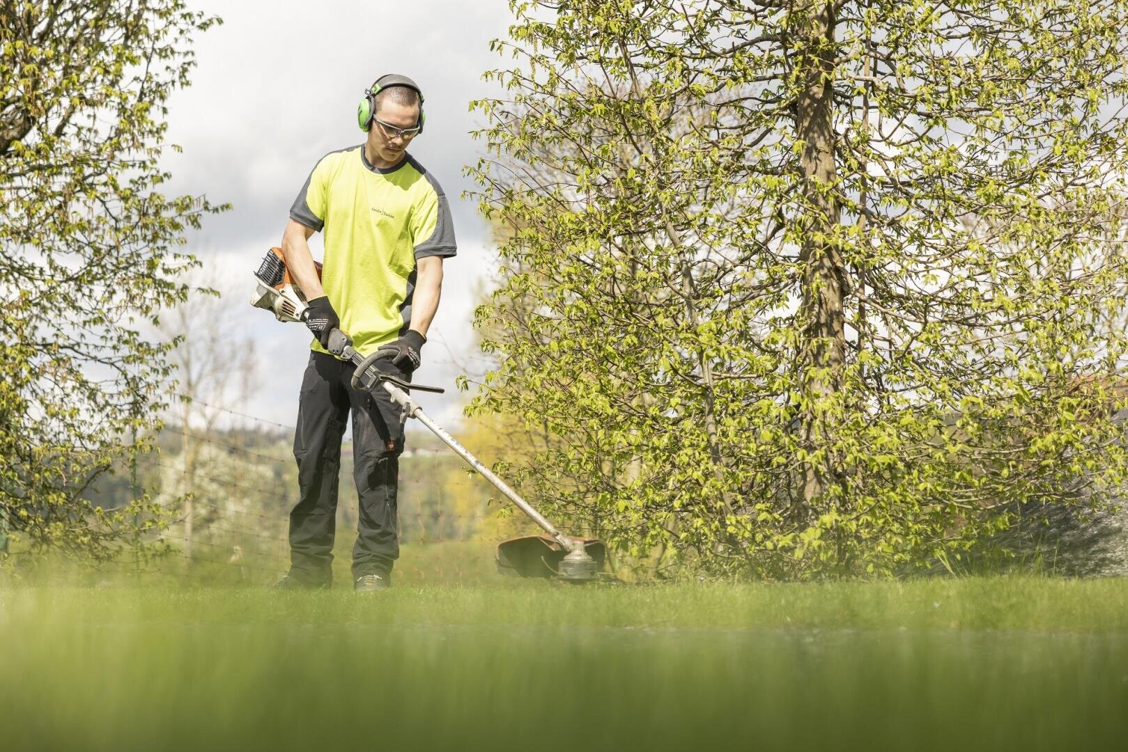 Eine Person, die eine Schutzbrille, einen Gehörschutz, Handschuhe und ein neongelbes Hemd trägt, schneidet an einem sonnigen Tag im Freien, umgeben von grünen Bäumen und Laub, mit einem Rasentrimmer Gras.