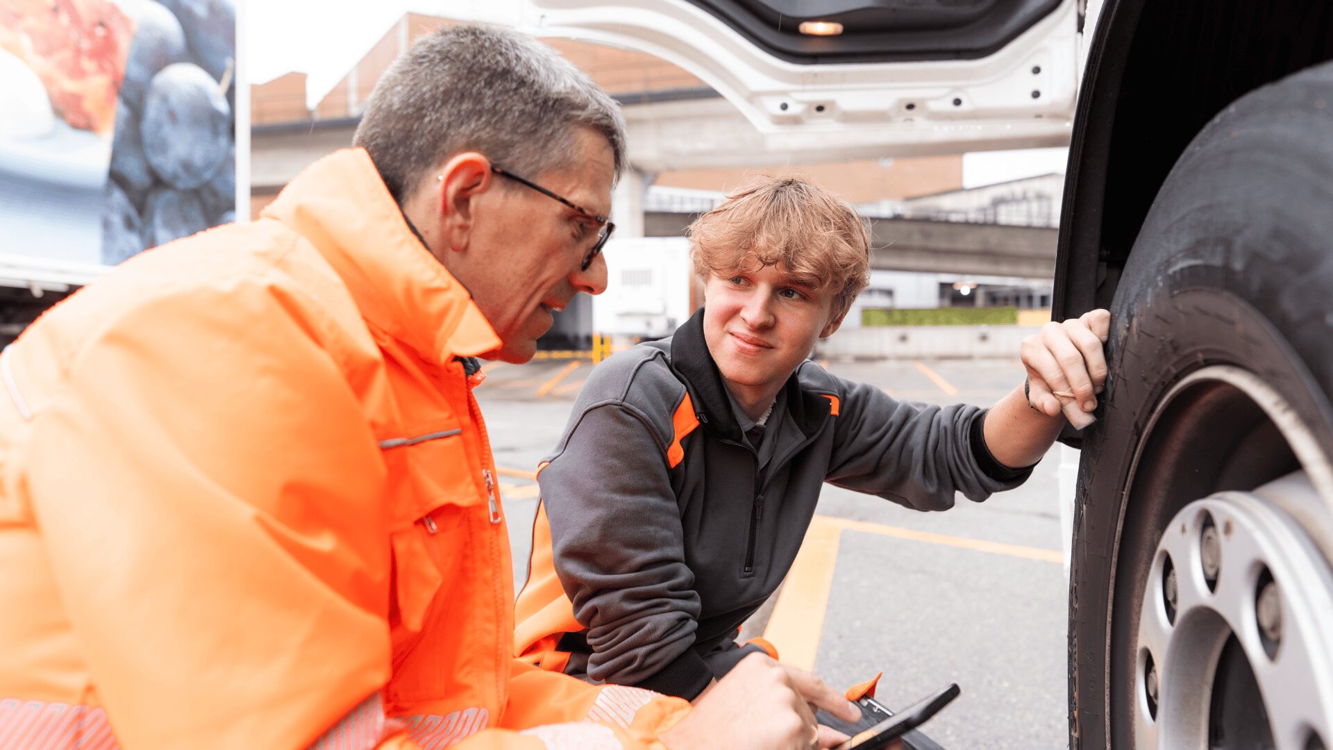Zwei Männer in Arbeitsuniformen untersuchen den Reifen eines großen Lastwagens. Ein älterer Mann in einer orangefarbenen Jacke hält ein Gerät, während ein jüngerer Mann ihn ansieht und den Reifen berührt. Sie scheinen sich über den Zustand des Reifens zu unterhalten.