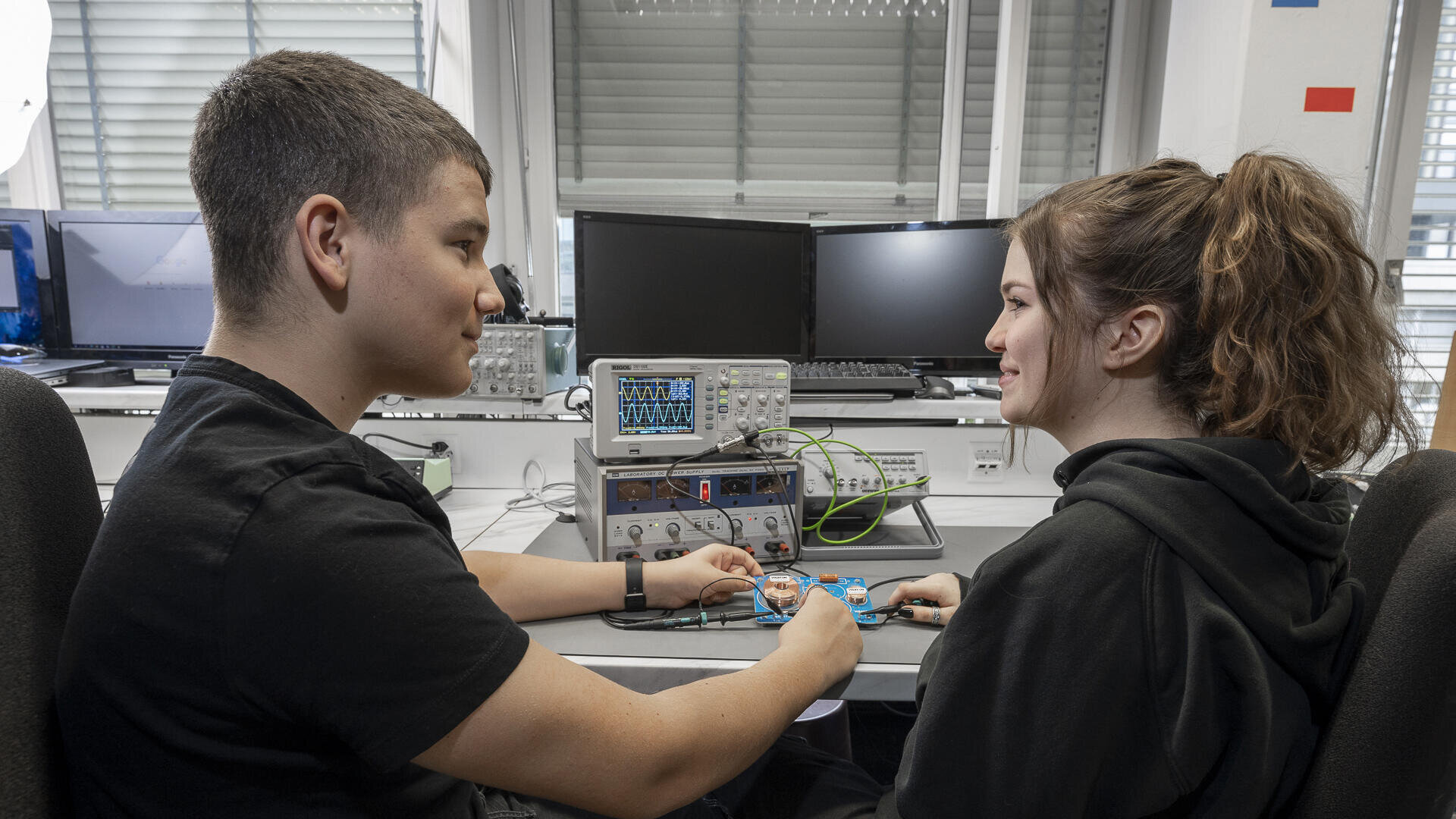 Zwei junge Leute sitzen sich in einem Labor gegenüber und halten elektronische Bauteile, die mit Drähten verbunden sind. Oszilloskope, Computer und andere Geräte stehen auf dem Schreibtisch hinter ihnen. Beide tragen schwarze Hemden und lächeln leicht.