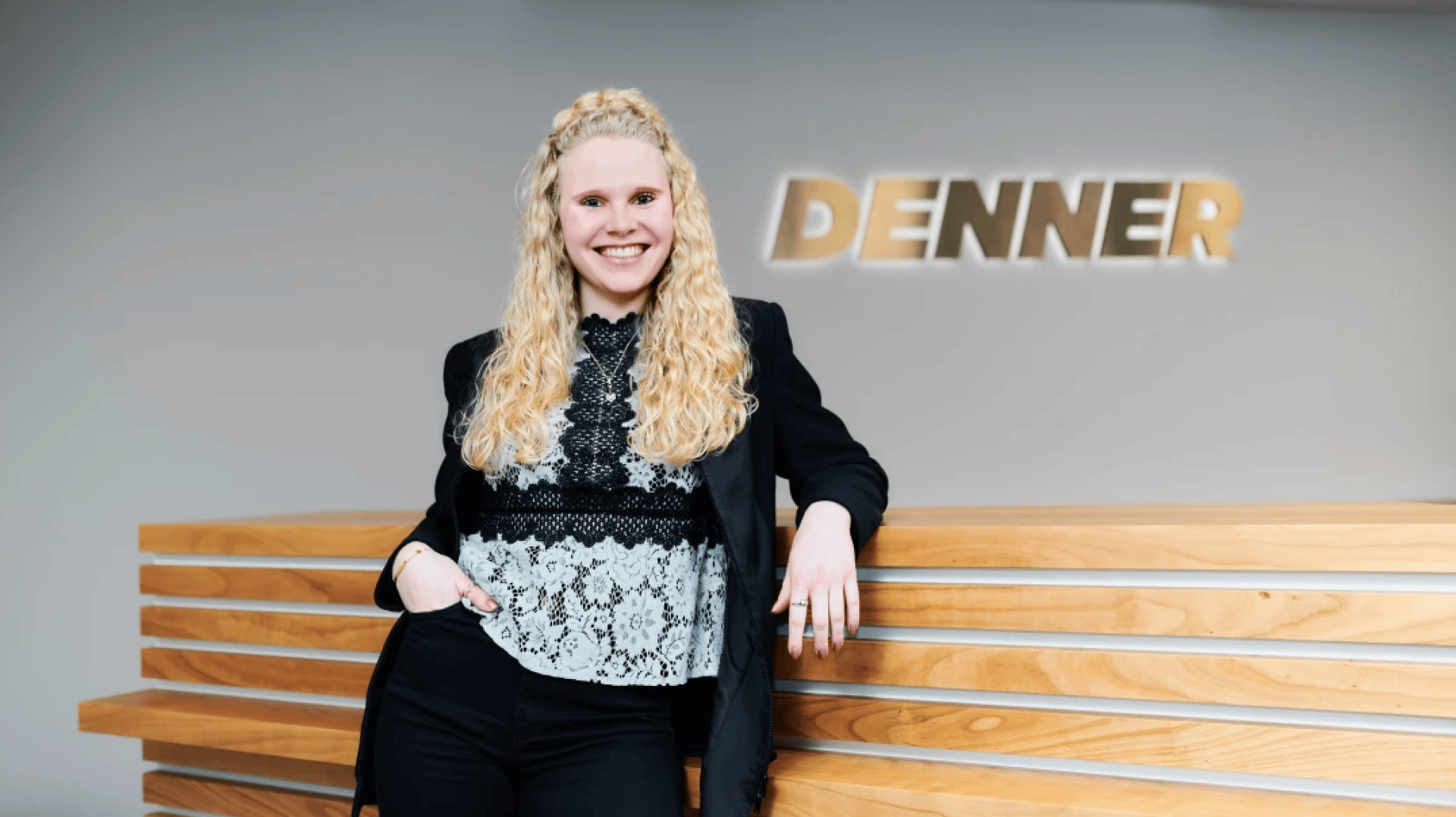 Eine lächelnde Frau mit langem, lockigem, blondem Haar, gekleidet in einen schwarzen Blazer und ein gemustertes Oberteil, steht selbstbewusst mit einem Arm auf dem hölzernen Empfangstresen gestützt. An der Wand hinter ihr ist das Wort DENNER zu lesen.