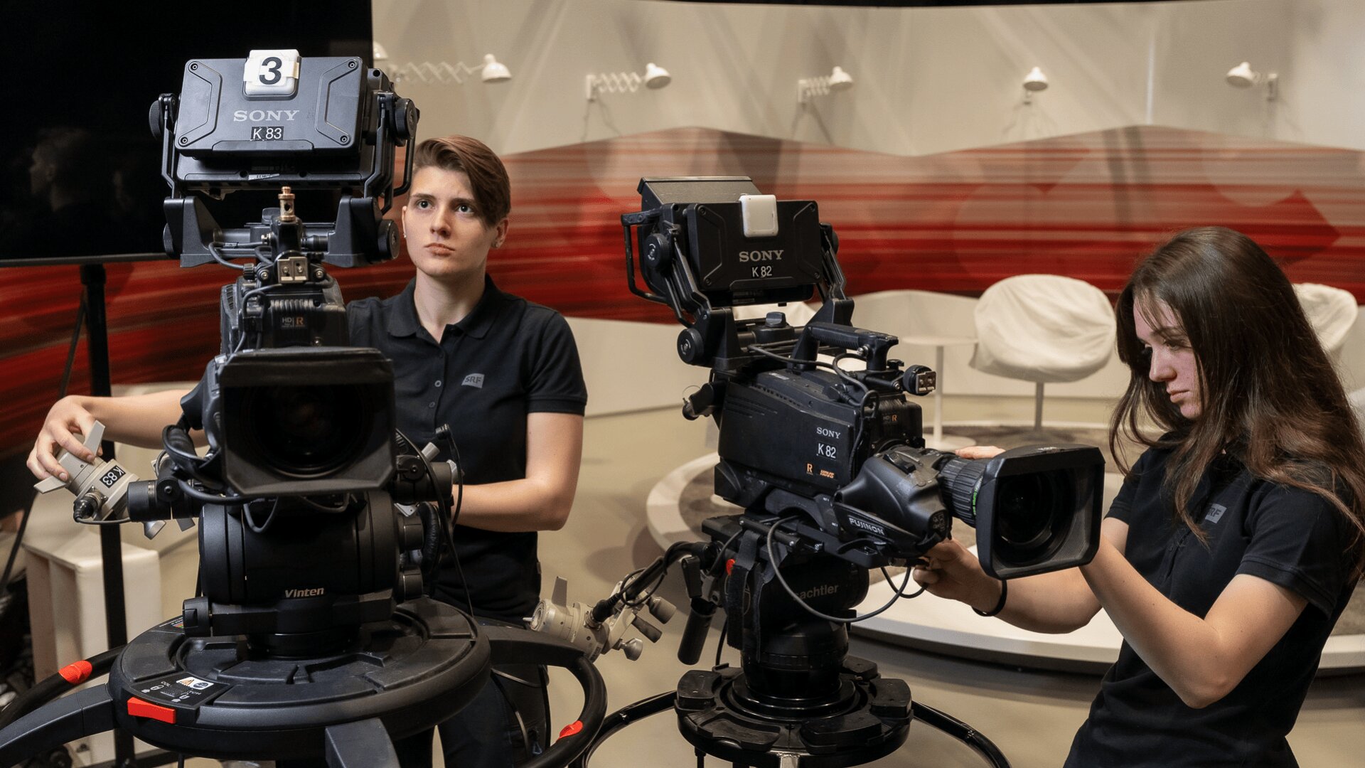Zwei Frauen in schwarzen Hemden bedienen große Videokameras in einem Studio mit rot-weißem Dekor und leeren weißen Stühlen im Hintergrund. Beide sind damit beschäftigt, ihre Ausrüstung einzustellen.