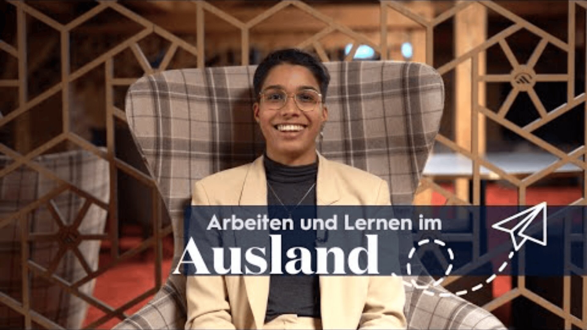 Eine Person in einem beigen Anzug und mit Brille sitzt lächelnd in einem karierten Sessel. Der überlagerte Text lautet: Arbeiten und Lernen im Ausland" mit einem Papierflugzeug als Grafik. Geometrische Holzmuster befinden sich im Hintergrund.