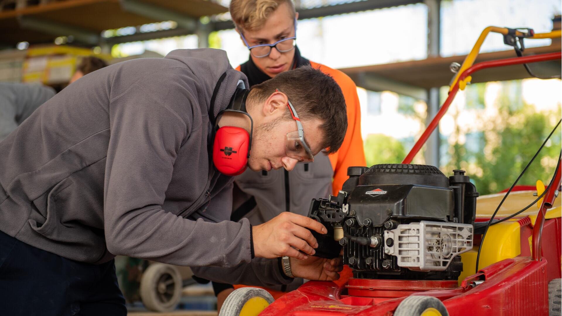 Zwei junge Leute arbeiten in einer Werkstatt an der Reparatur oder Inspektion eines roten Rasenmähermotors. Eine Person untersucht den Motor genau, während sie eine Schutzbrille und einen Gehörschutz trägt; die andere schaut aufmerksam zu.