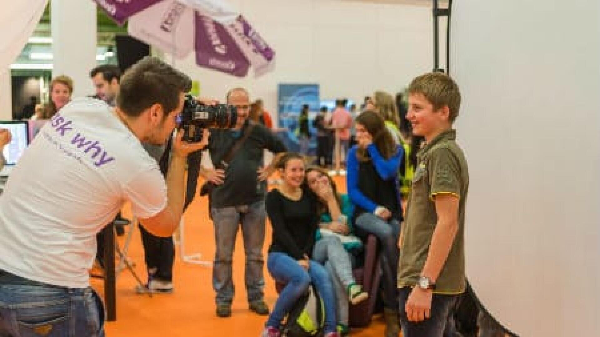 Ein Fotograf macht ein Foto von einem lächelnden Jungen, der vor einem weißen Hintergrund posiert, während eine Gruppe von Menschen im Hintergrund bei einer Indoor-Veranstaltung zusieht und lächelt.