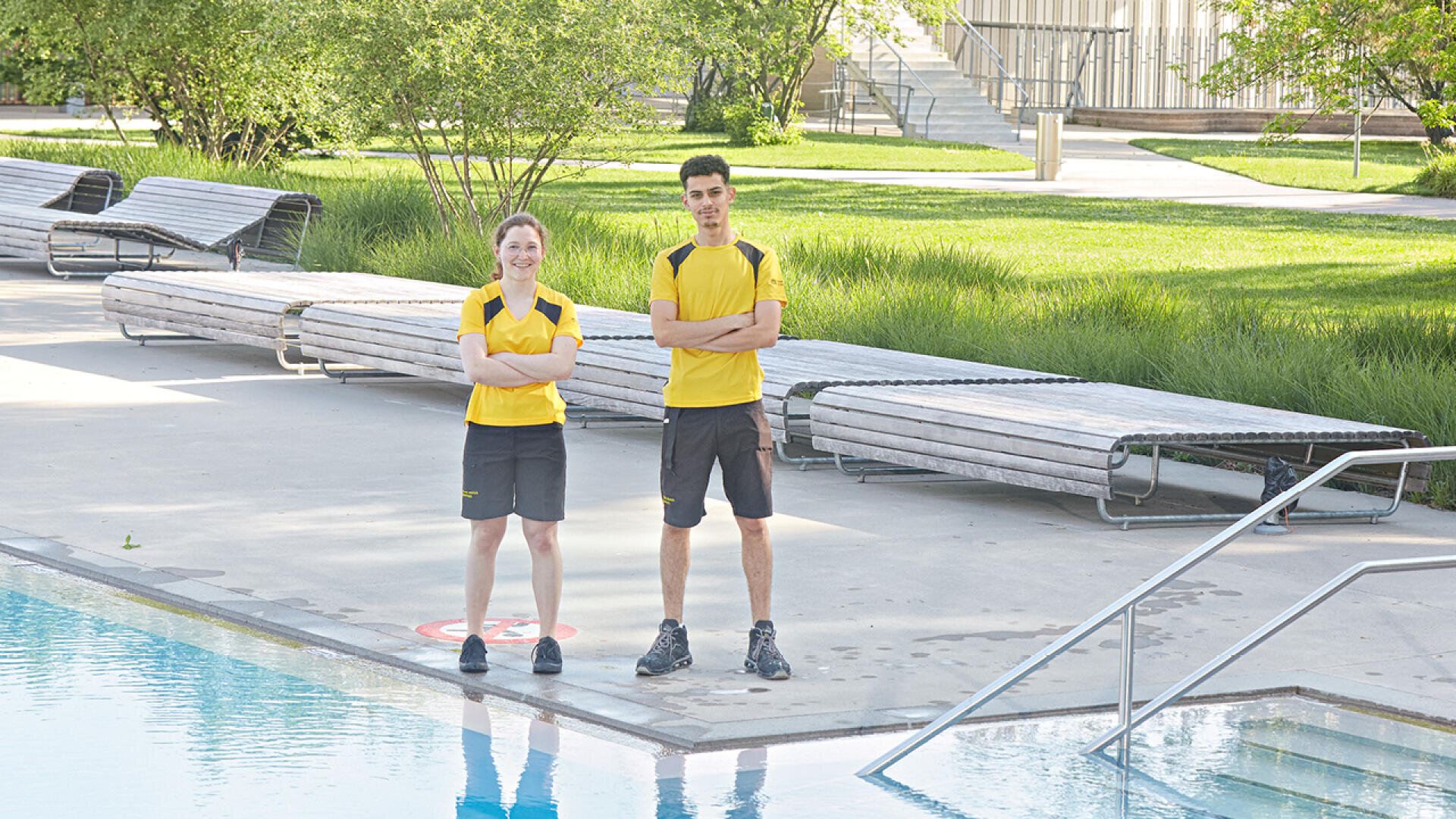 Zwei Rettungsschwimmer in gelben Hemden und dunklen Shorts stehen mit verschränkten Armen am Rande eines Freibads, mit Liegestühlen und grünen Bäumen im Hintergrund.