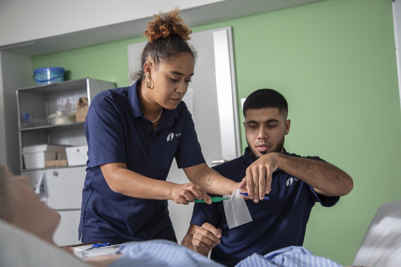 Eine Frau in einer marineblauen Uniform hilft einem Mann, ebenfalls in einer marineblauen Uniform, den Arm zu verbinden, während sie sich um einen Patienten in einer medizinischen Einrichtung mit grünen Wänden und medizinischen Geräten im Hintergrund kümmern.