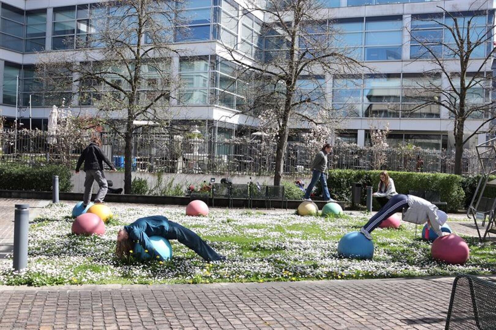 Menschen trainieren auf bunten Yogabällen, die auf einer Wiese mit weißen Blumen verstreut sind, umgeben von modernen Glasgebäuden. Einige dehnen sich oder beugen sich auf den Bällen nach hinten. Die Szene ist hell und im Freien.