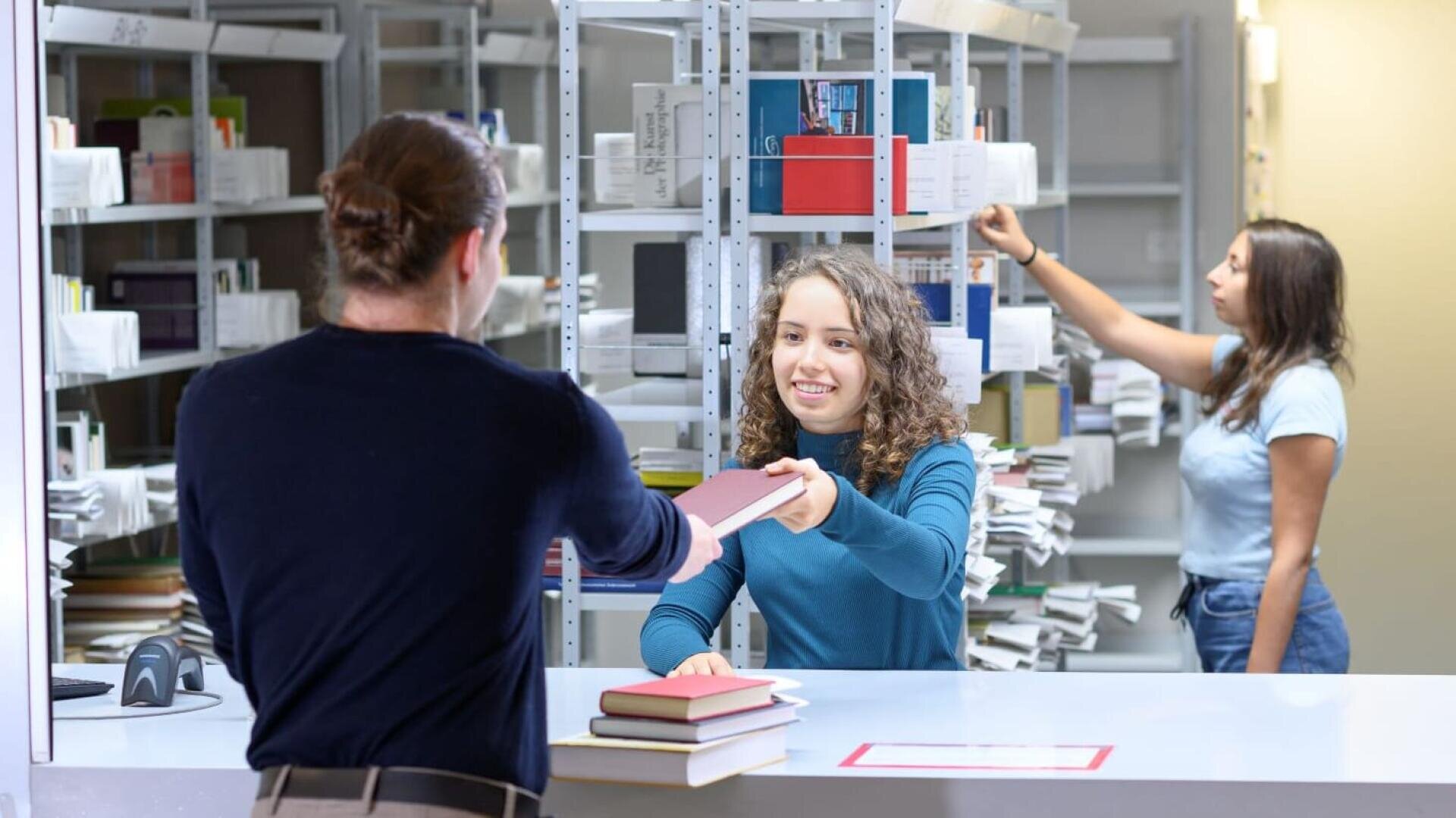 Eine junge Frau hinter einem Bibliothekstresen reicht einem Mann ein Buch, während eine andere Frau im Hintergrund in den Bücherregalen stöbert. Zu sehen sind Bücherstapel und Bibliotheksregale.