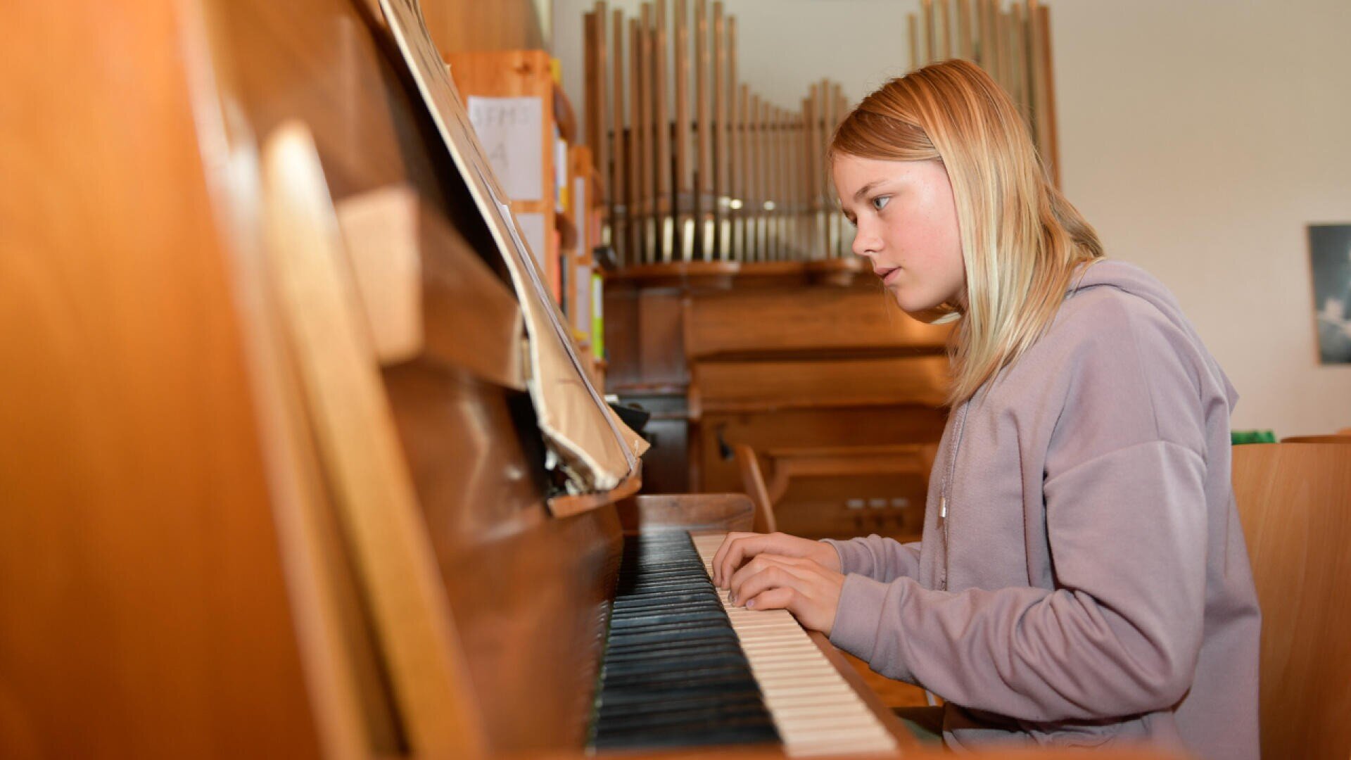 Eine junge Person mit blondem Haar und lavendelfarbenem Kapuzenpulli spielt auf einem Klavier in einem Raum mit Holzmöbeln und Orgelpfeifen im Hintergrund.