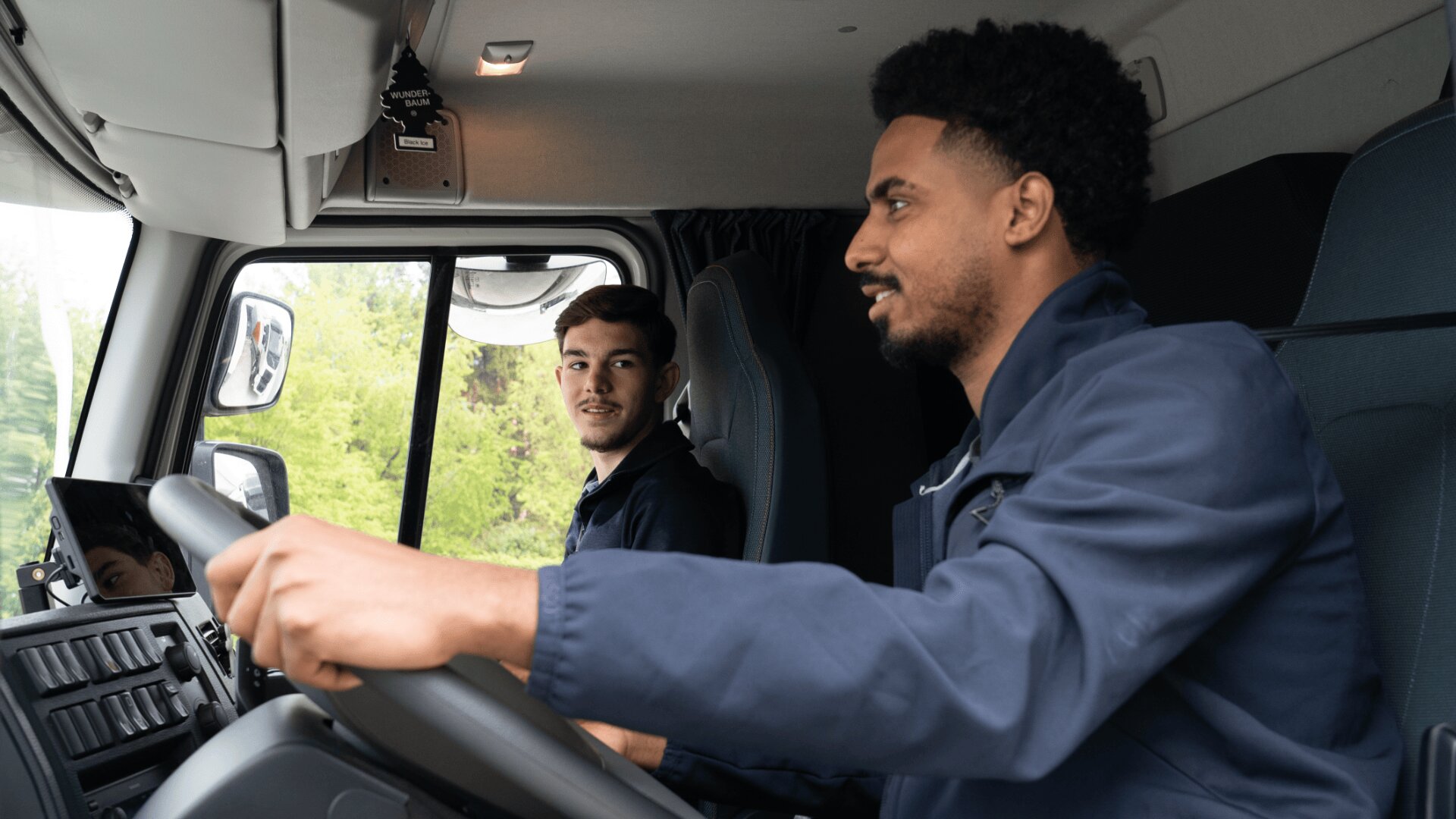 Zwei Männer in dunklen Uniformen sitzen im Führerhaus eines Lastwagens; der eine fährt und lächelt, während der andere, der neben ihm sitzt, ihn anschaut, beide wirken konzentriert und engagiert.