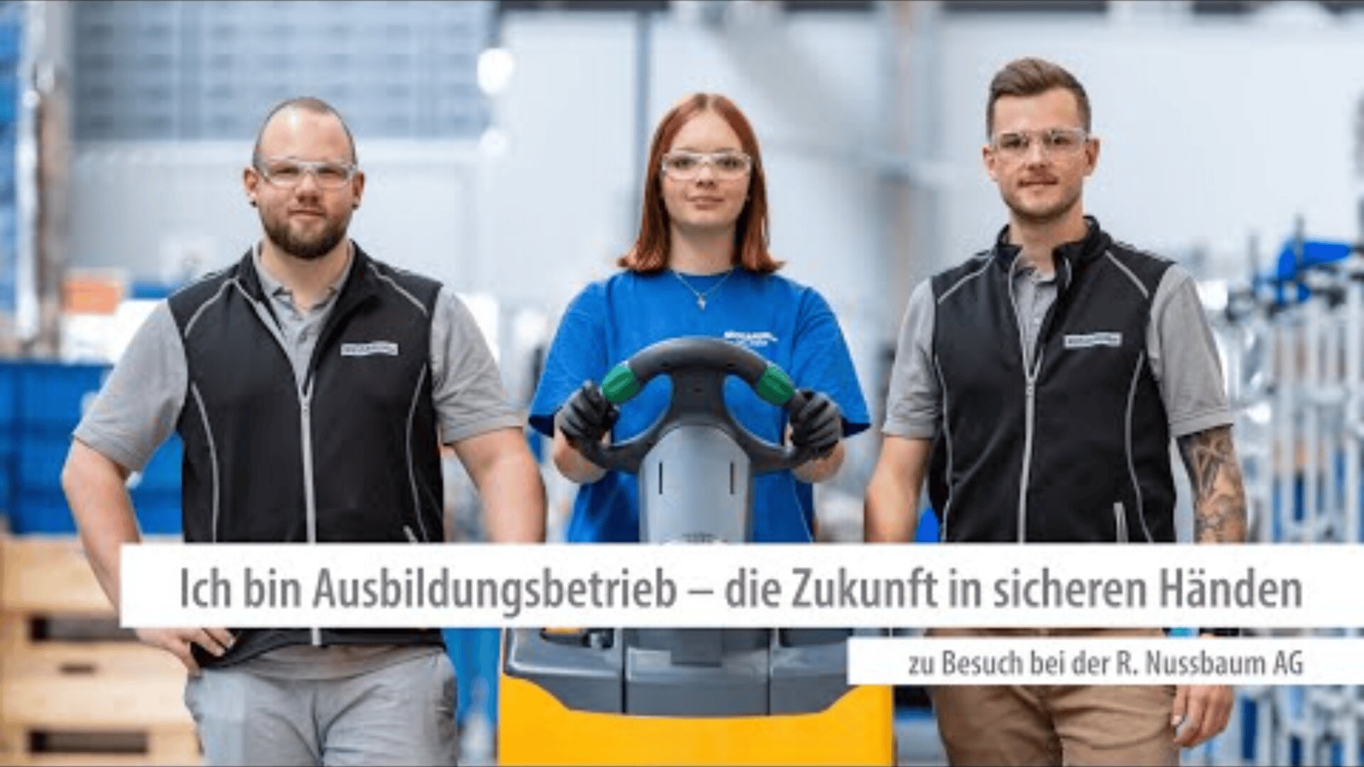 Drei junge Erwachsene mit Schutzbrillen und Arbeitsuniformen stehen in einer Industrieumgebung zusammen. Die Frau in der Mitte hält eine Ausrüstung. Der Text auf Deutsch lautet: Ich bin Ausbildungsbetrieb - die Zukunft in sicheren Händen.