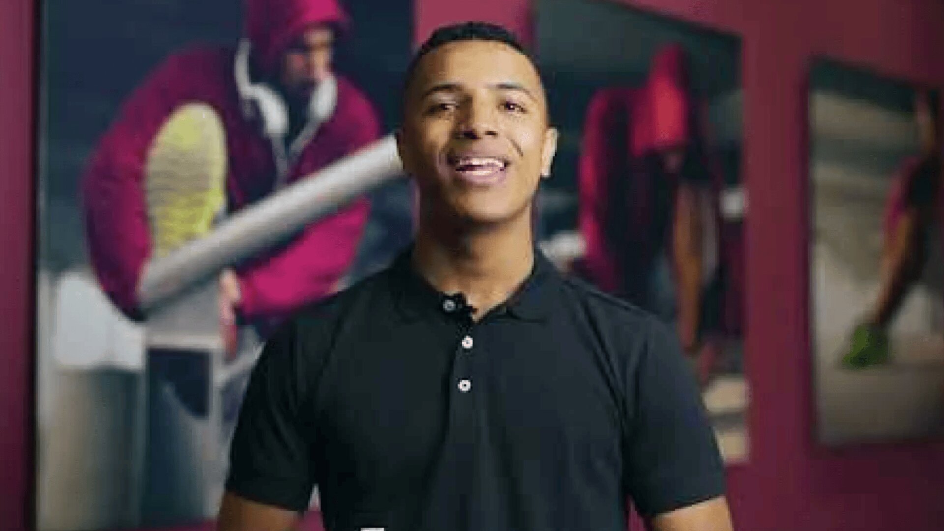 Eine Person in einem schwarzen Poloshirt steht in einem Haus und lächelt in die Kamera. Im Hintergrund sind unscharfe Bilder von Sportlern in roten Kapuzenpullovern zu sehen, die wahrscheinlich laufen oder sich auf ein Rennen vorbereiten.
