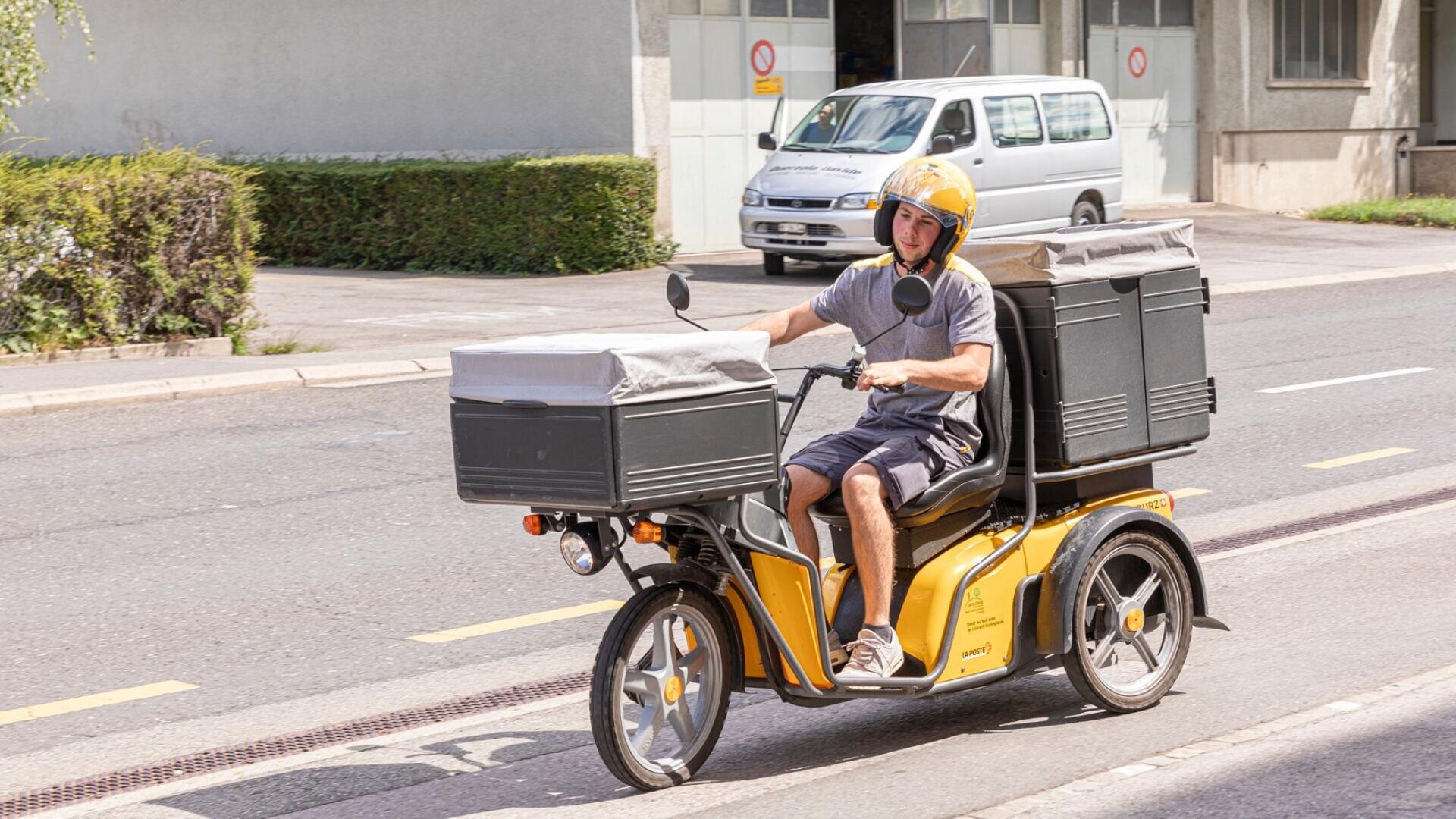 Eine Person, die einen gelben Helm trägt, fährt tagsüber auf einer städtischen Straße einen gelben dreirädrigen Lieferroller mit großen schwarzen Aufbewahrungsboxen.